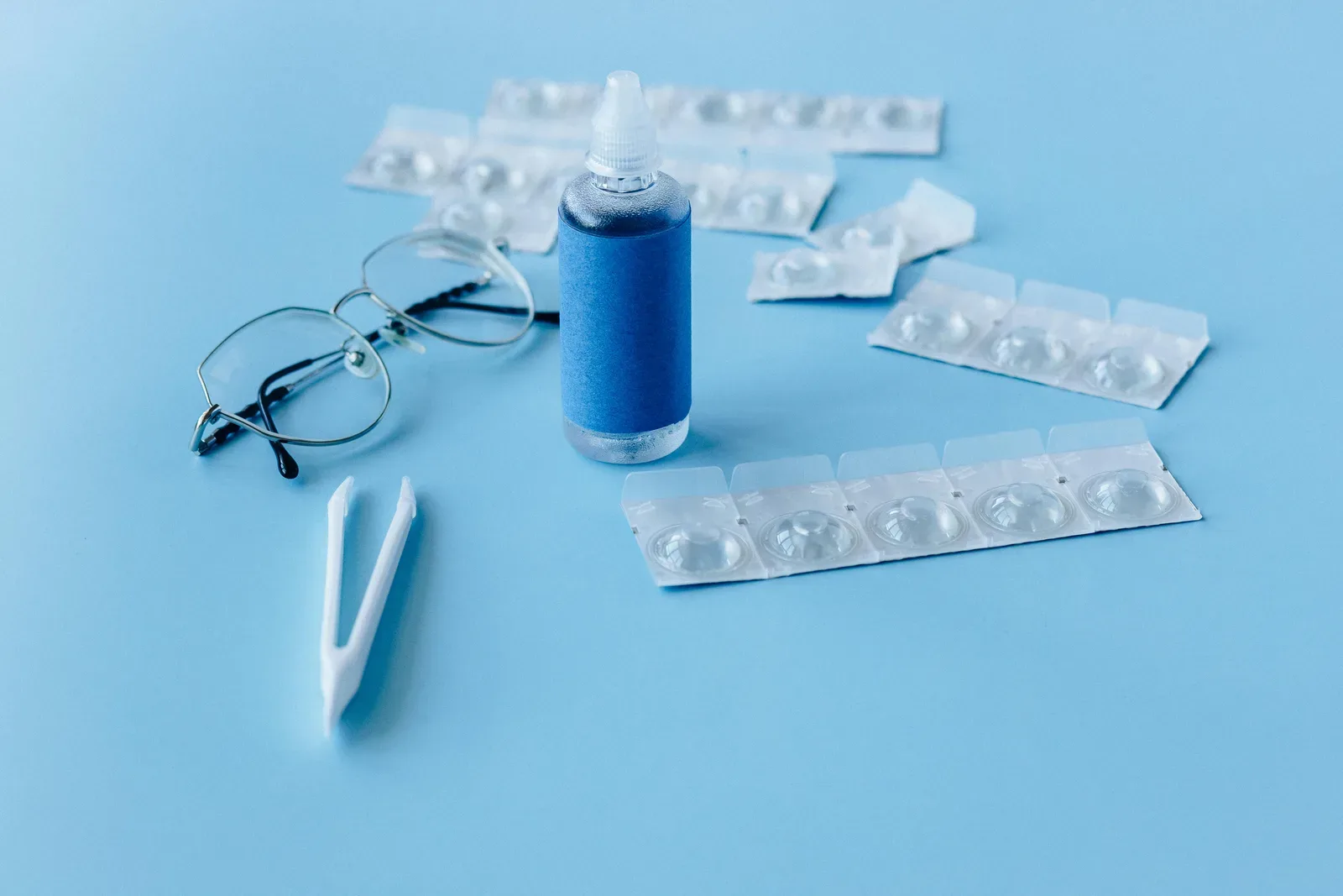 Close-up of contact lens solution bottle with eyeglasses and tweezers on blue backdrop.