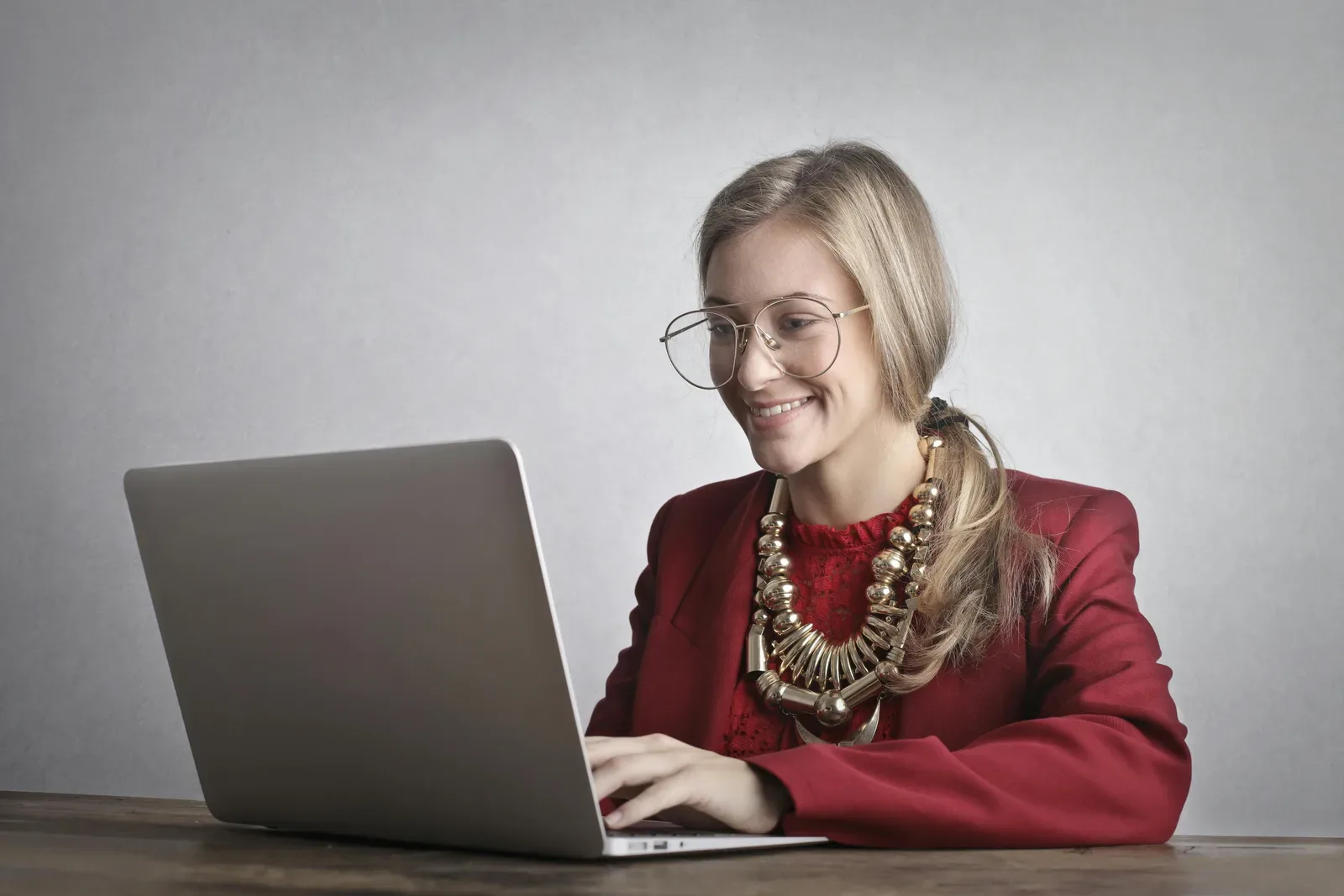A blonde woman in a red coat smiles while using a laptop indoors.