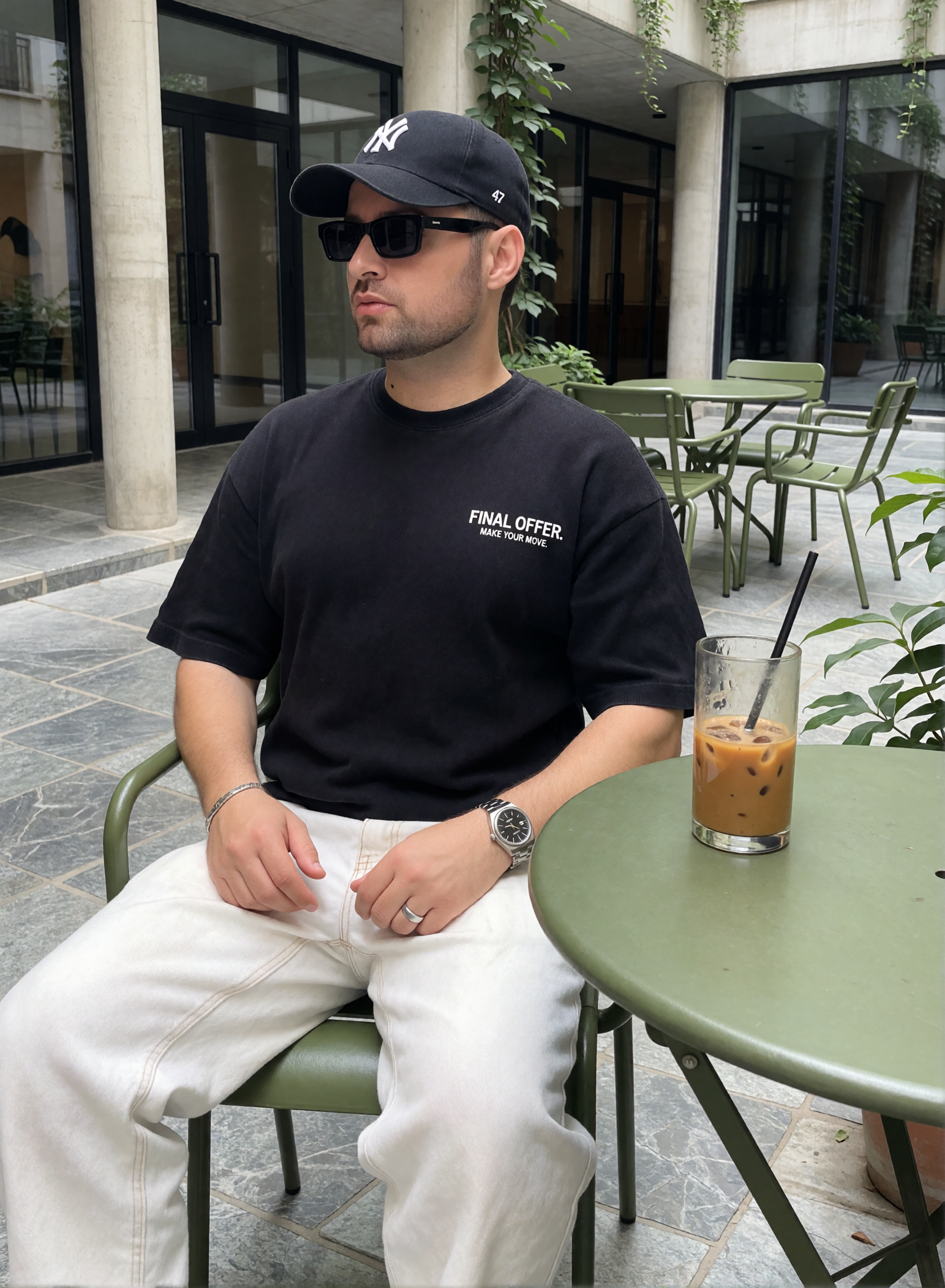 Digitaler Zwilling von Bruno Dias mit schwarzem T-Shirt, weißer Hose, Sonnenbrille und Cap beim Eiskaffee im begrünten Innenhof.