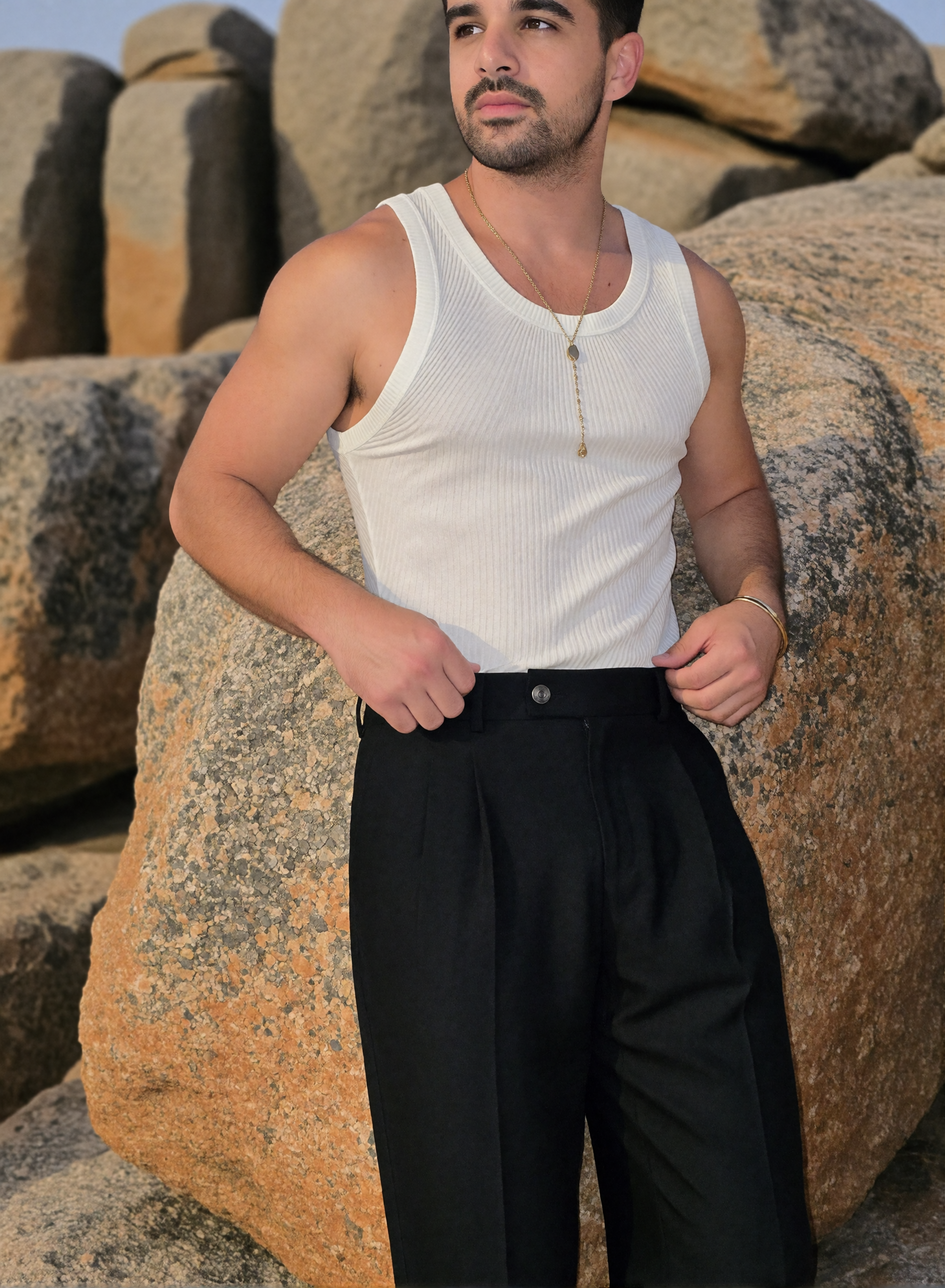 Digitaler Zwilling Mousaab Ainouche im weißen gerippten Tanktop und schwarzer Bundfaltenhose, mit goldfarbener Halskette vor sonnenbeschienenen Felsen.