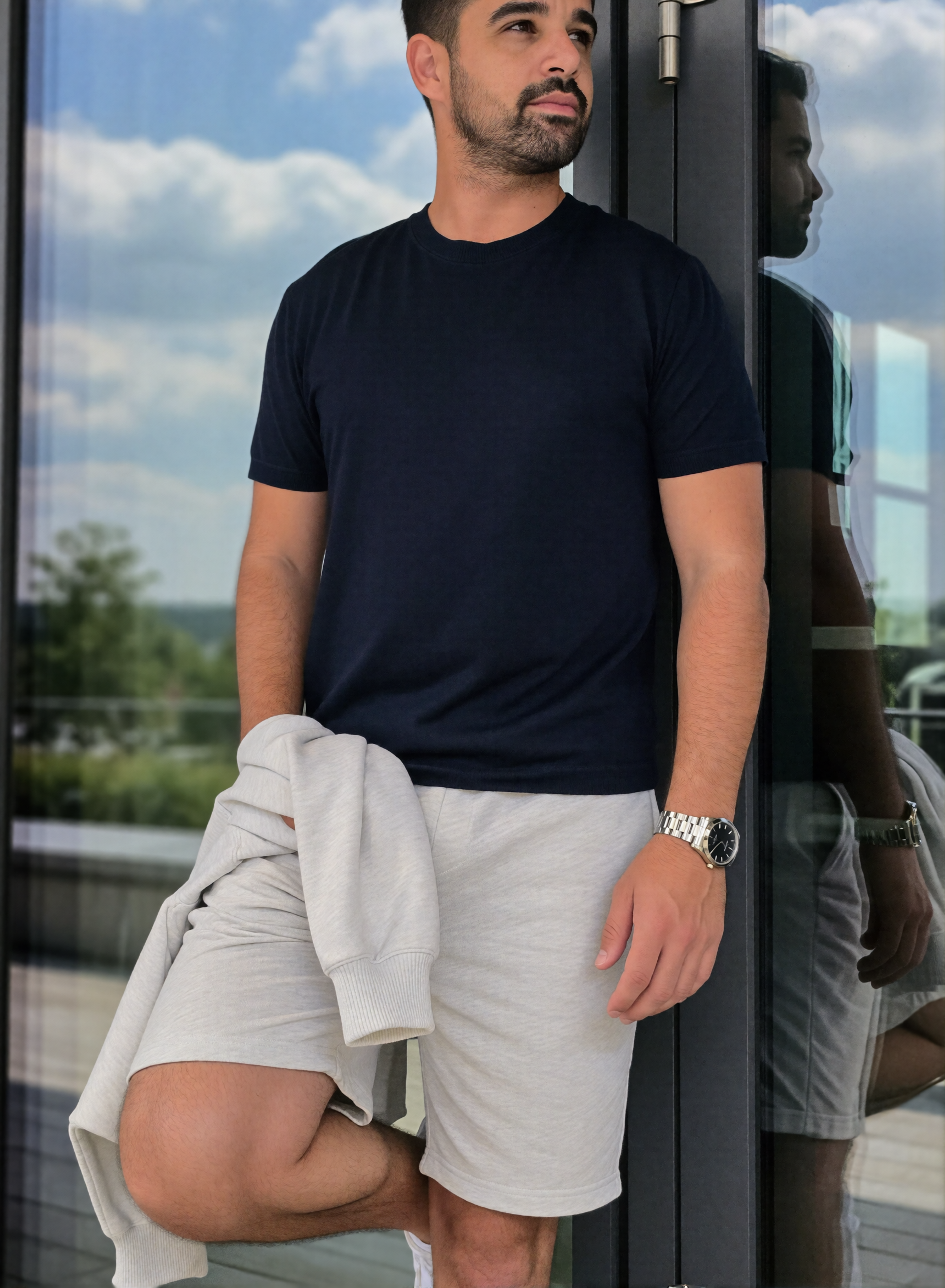  Digitaler Zwilling Mousaab Ainouche in dunkelblauem T-Shirt und hellgrauen Shorts, mit Sweatshirt und silberner Armbanduhr, an einer spiegelnden Glasfront.