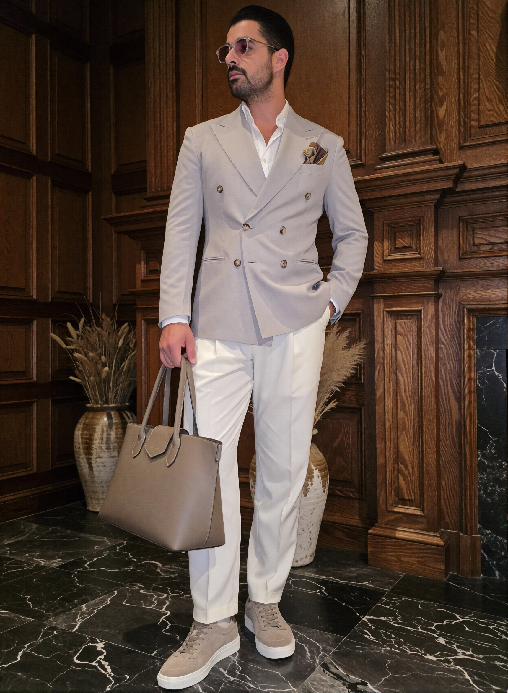 Digitaler Zwilling Mousaab Ainouche in hellgrauem Zweireiher, weißer Hose und beigen Sneakern, mit Sonnenbrille und taupefarbener Tasche vor dunkler Holzvertäfelung.