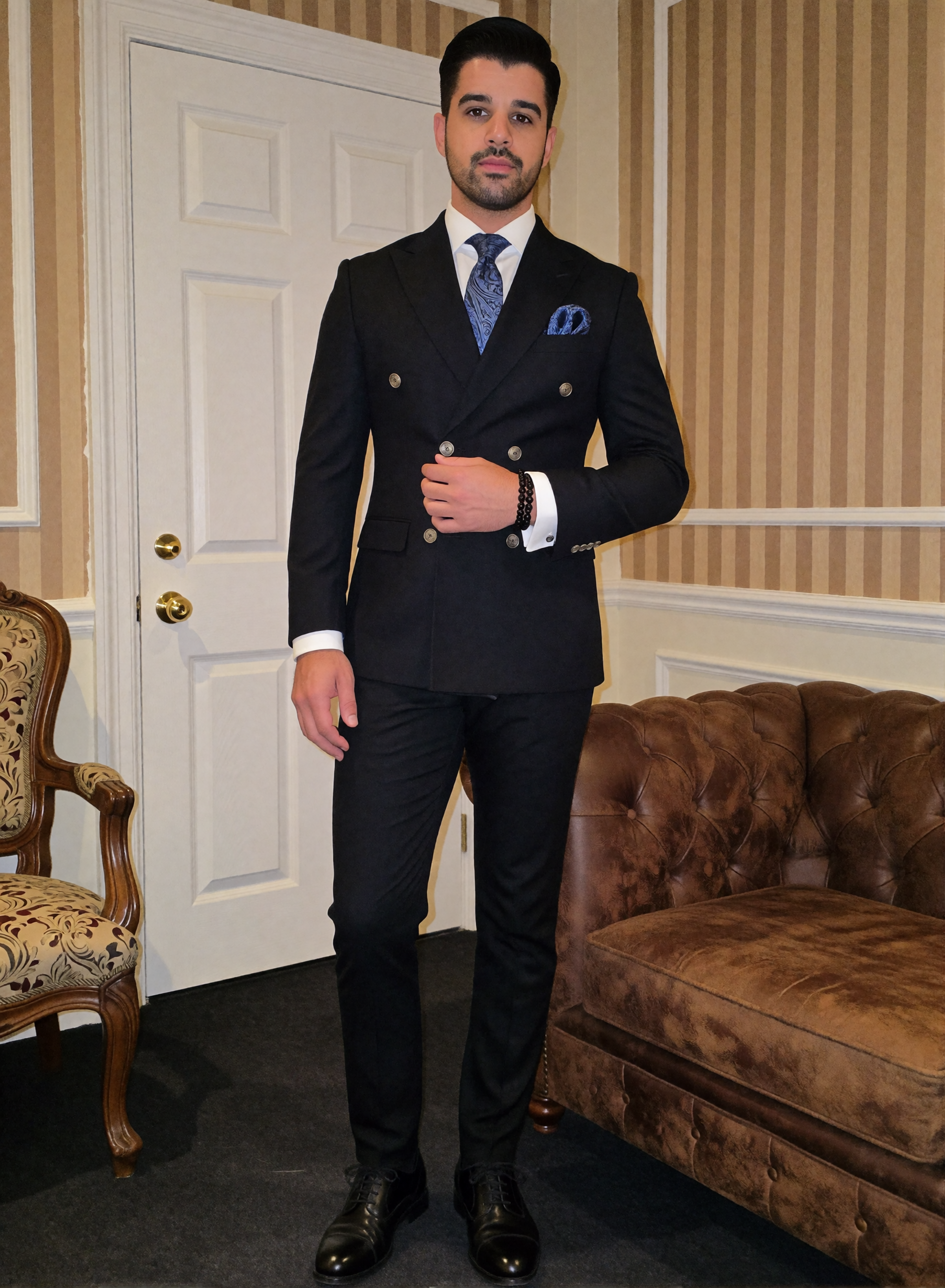 Digitaler Zwilling Mousaab Ainouche im dunklen Zweireiher mit weißem Hemd, blauer Krawatte und Einstecktuch, neben einem braunen Chesterfield-Sofa.