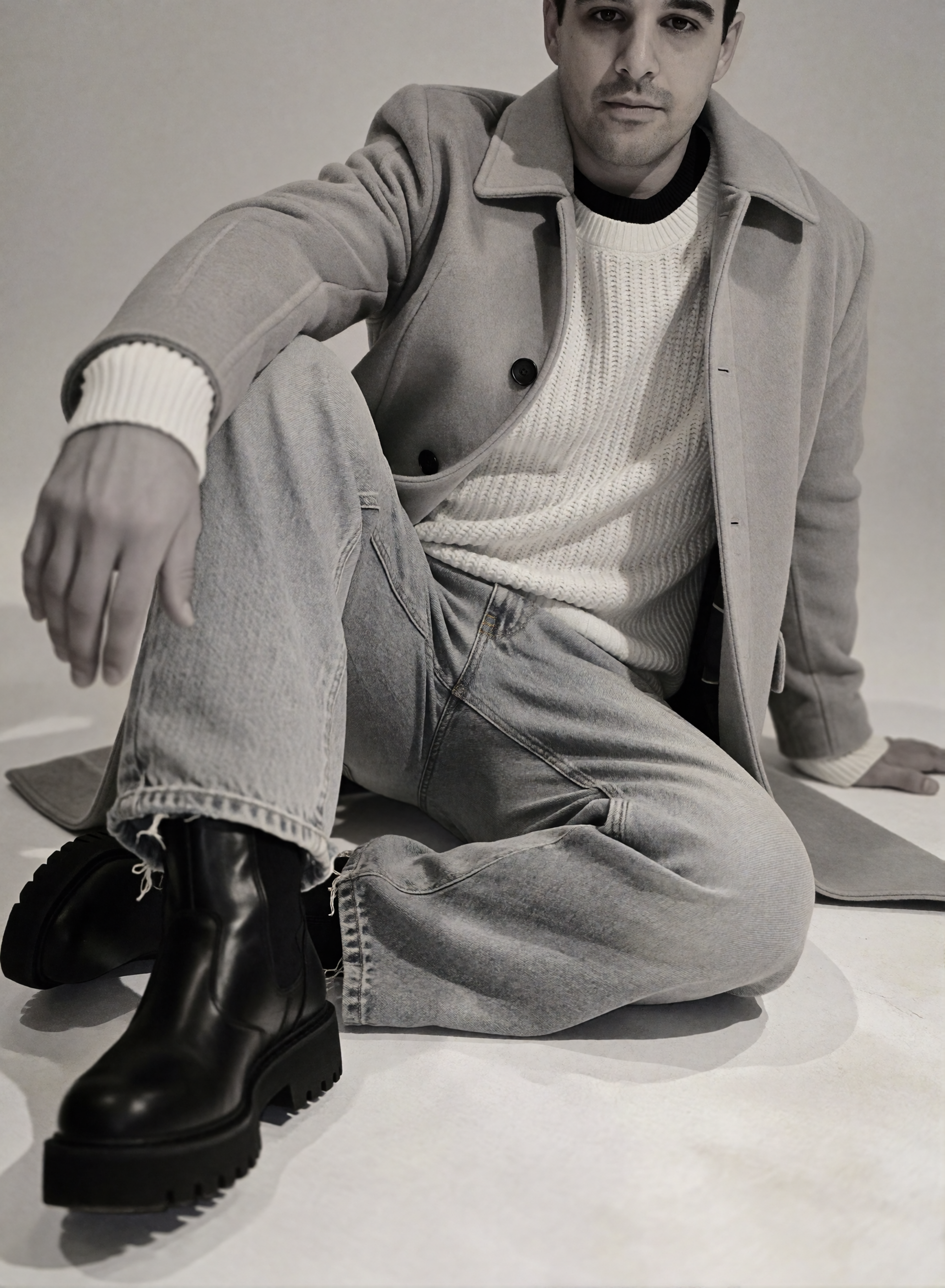 Digitaler Zwilling Mousaab Ainouche sitzt im hellen Mantel, groben Strickpullover, heller Jeans und schwarzen Chelsea-Boots auf dem Studioboden, in Schwarz-Weiß.