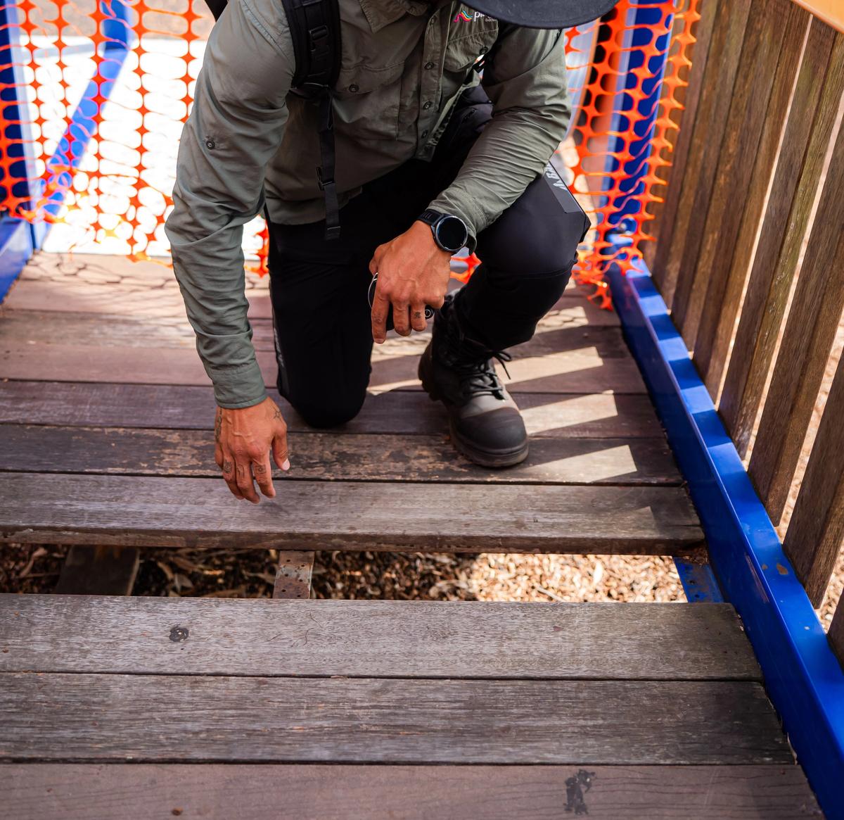 A Play Force inspector crouching on play equipment to inspect damage. The wooden walkway has planks missing, causing a significant hazard.