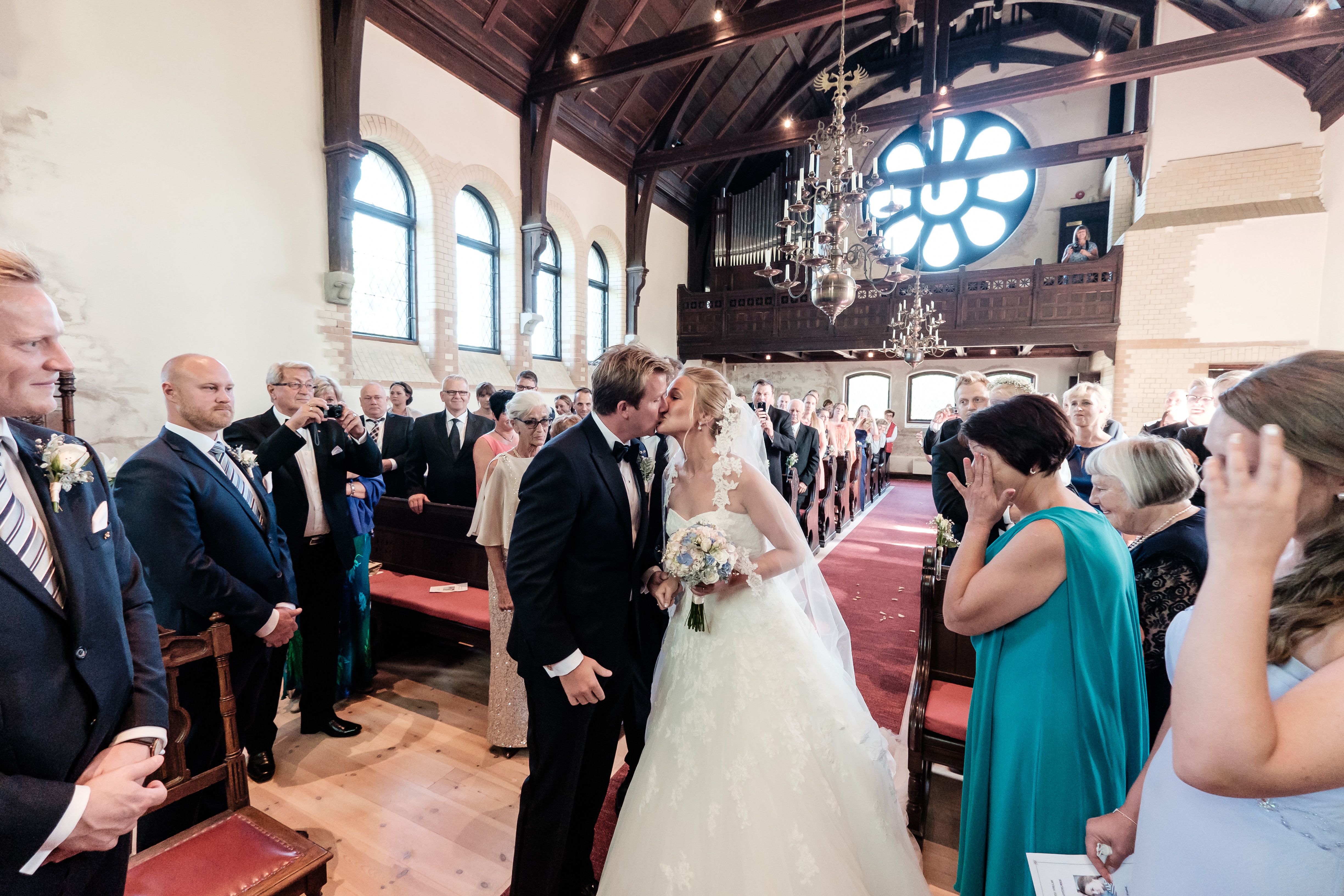 Brudepar kysser under vielsen i en vakker svensk kirke omgitt av familie og venner, med klassisk nordisk interiør og stemningsfull belysning.