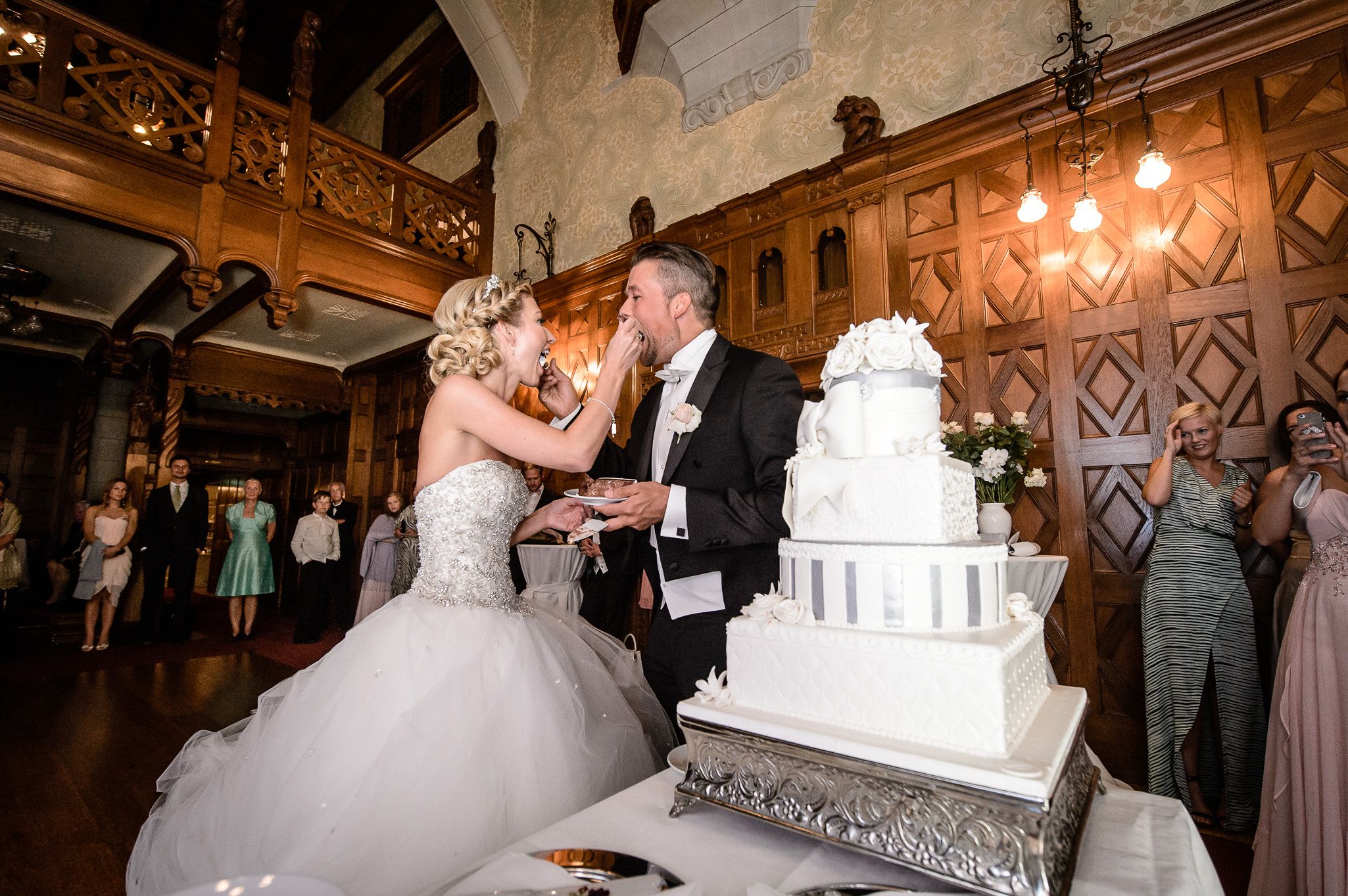 Brudepar deler bryllupskake på et svensk slott under feiringen, omgitt av gjester og elegant interiør.