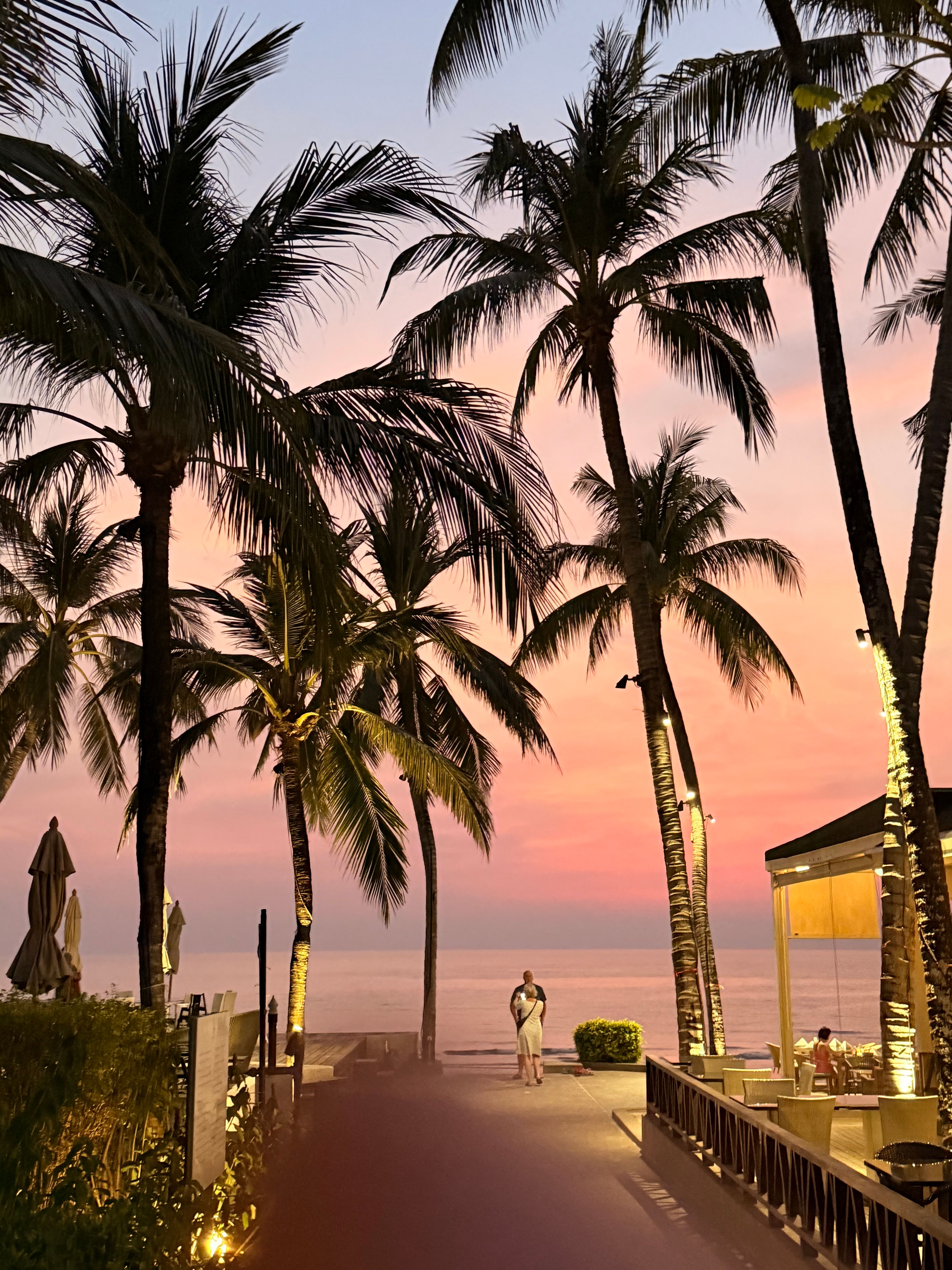 Par står under palmer i solnedgang ved stranden i Thailand, omgitt av rosa himmel og hav.