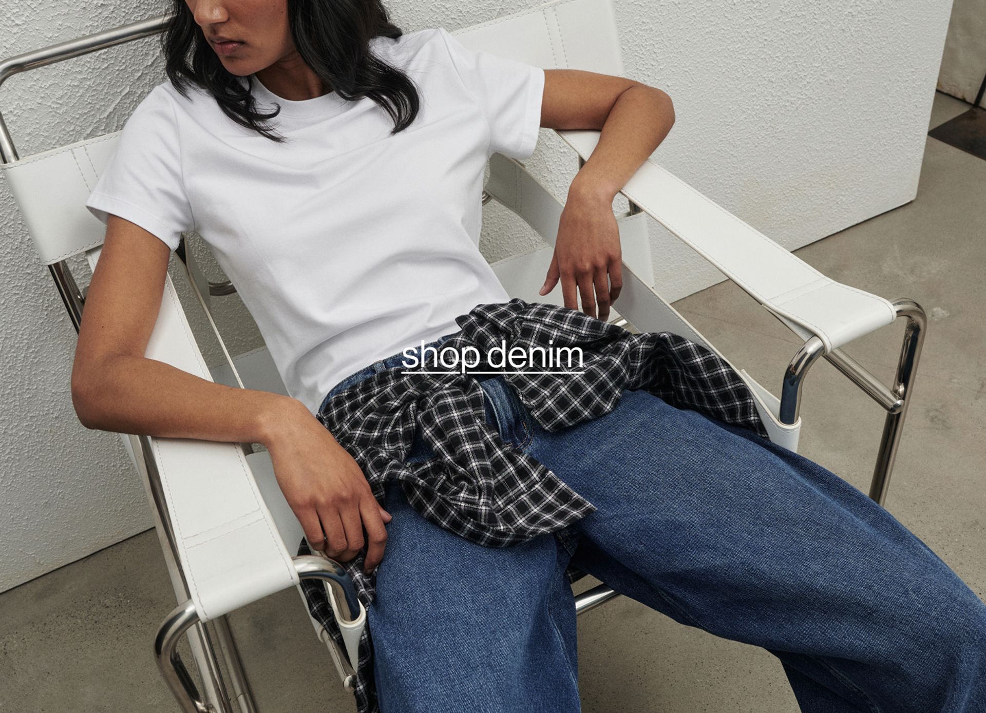 Model wearing a white tee and wide-leg blue jeans, sitting in a white chair against a light concrete wall.