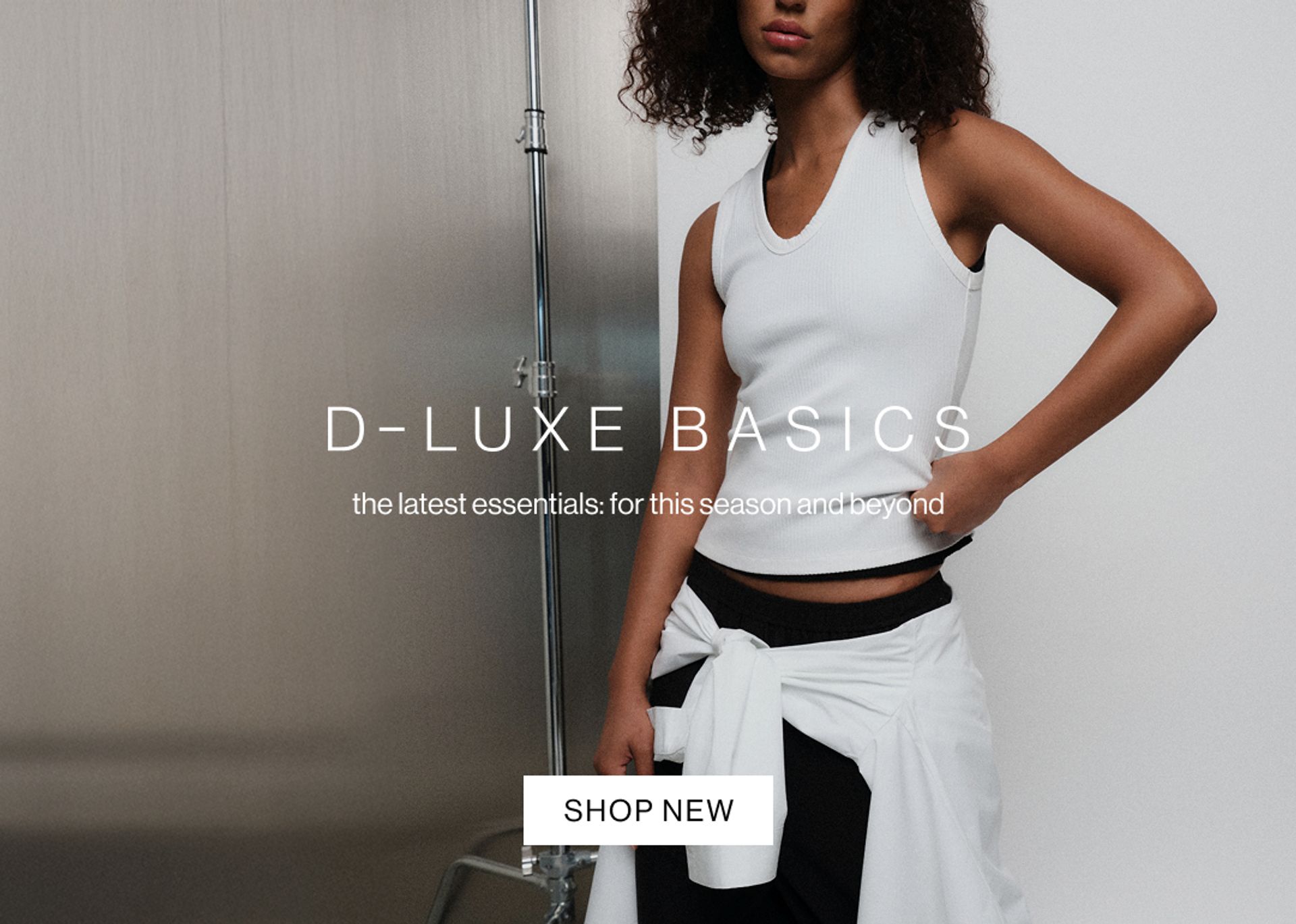 Model in a white tank and black pant with white shirt wrapped around waist against a modern metallic wall with “D-Luxe Basics” headline.