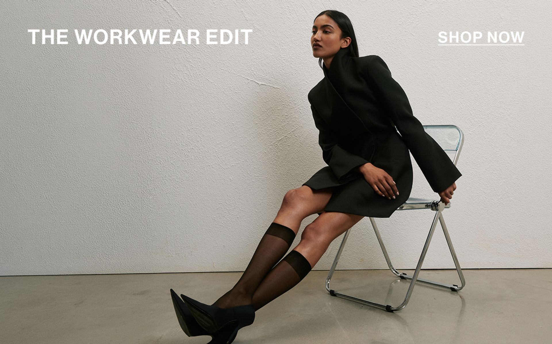 Model sitting on a chair wearing a black high neck blazer dress. SHOP THE WORKWEAR EDIT.