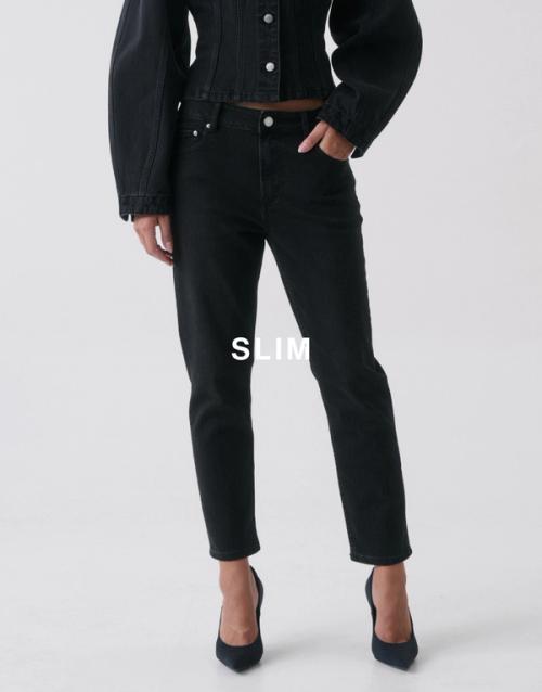 Jess Hart standing in black slim-fit jeans with a cropped black jacket, hand in pocket. SHOP SLIM JEANS.