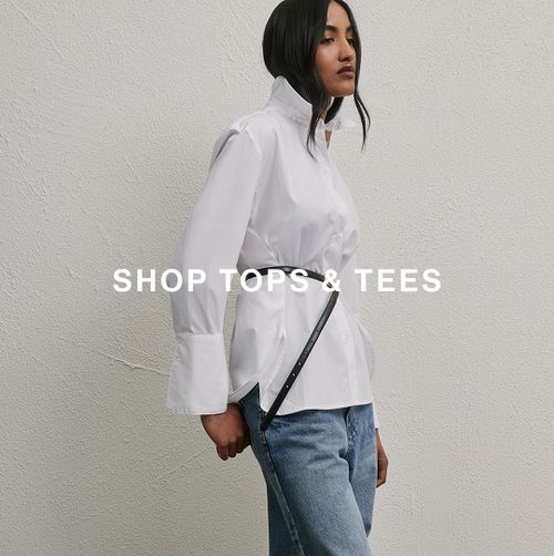 Model in a white blouse and jeans standing against a neutral wall – Shop Tops.