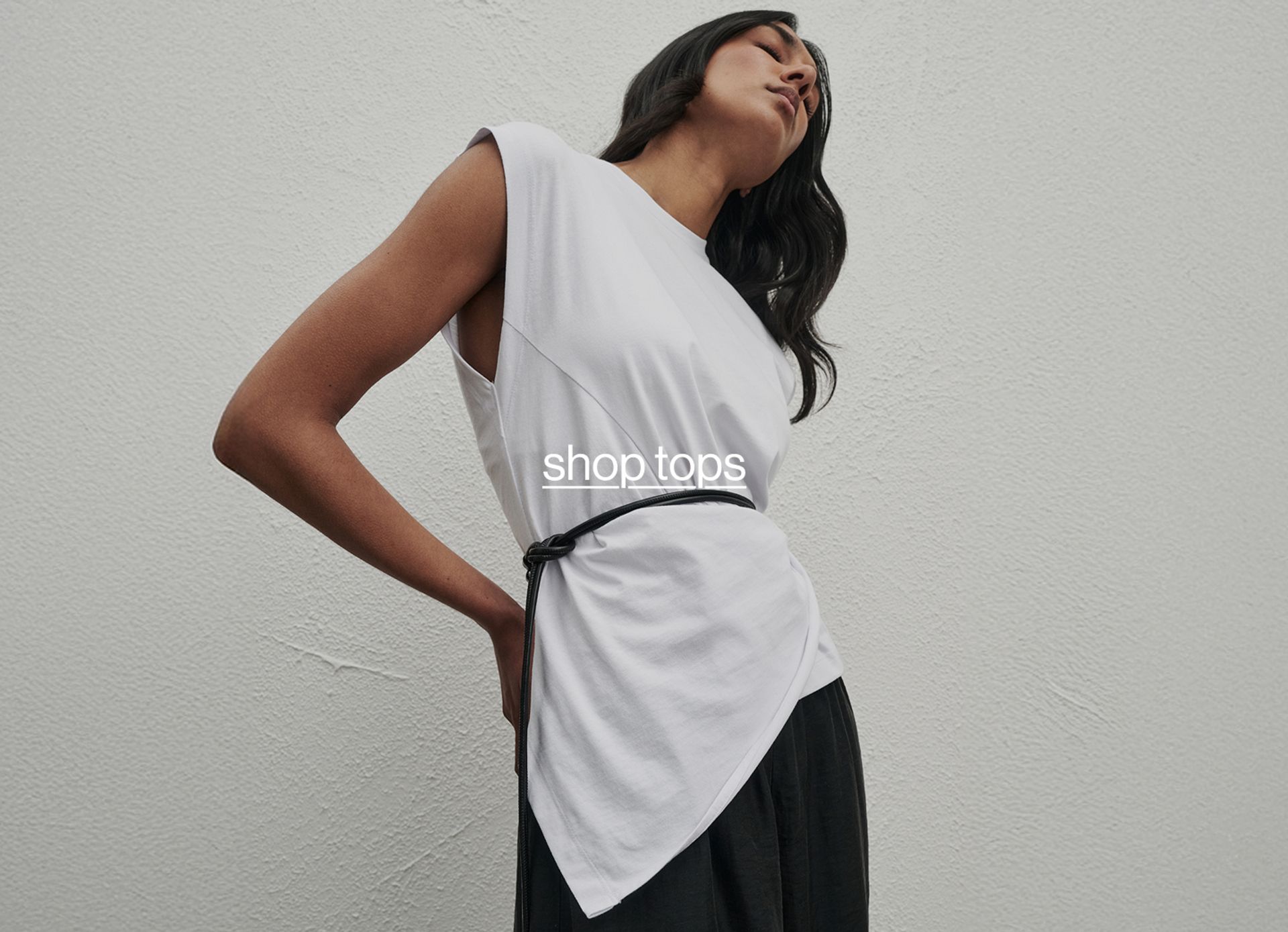 Model in a white sleeveless top layered over black, posing side-on against a textured neutral wall.