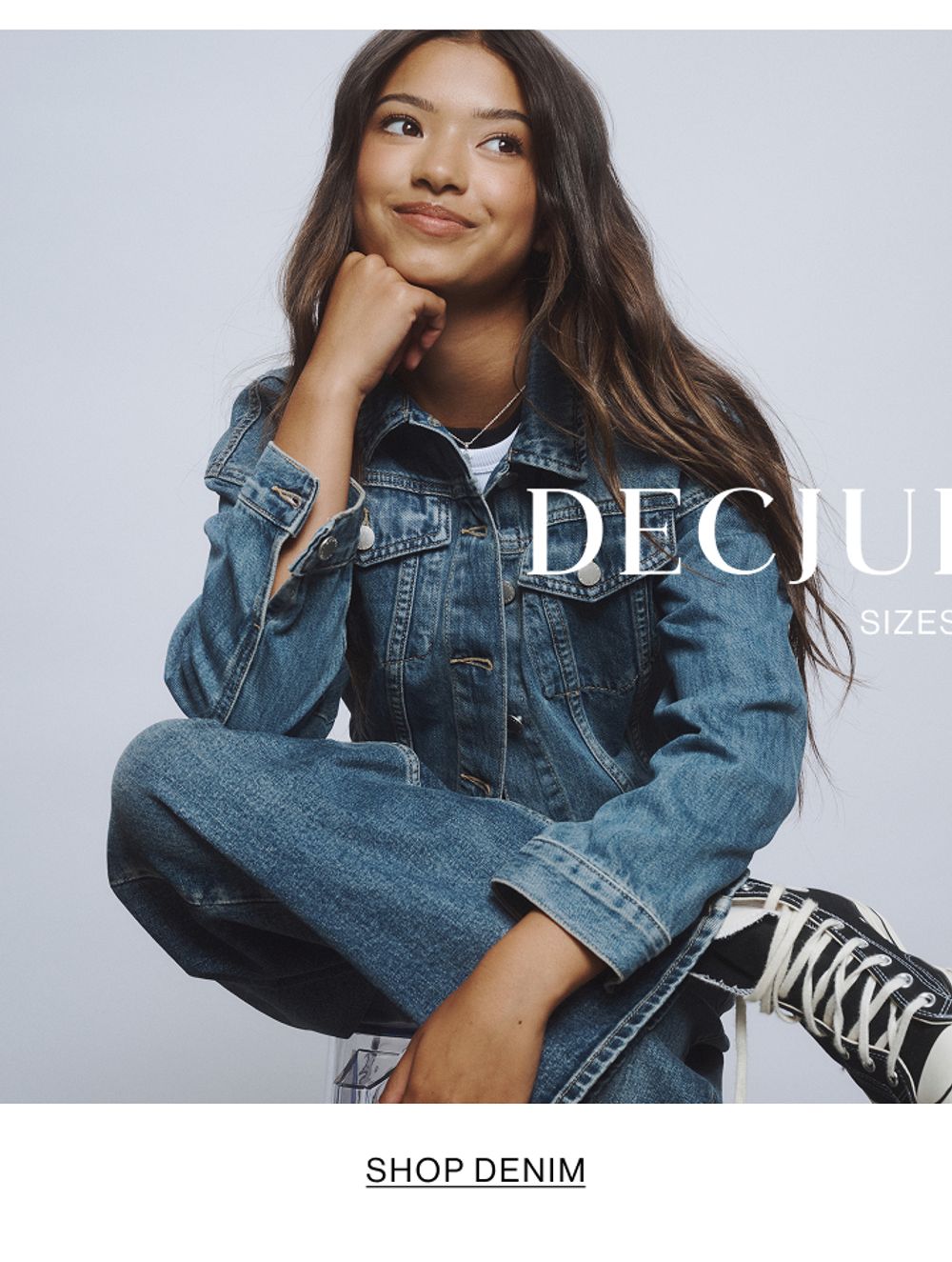 Young girl sitting cross-legged wearing a denim jacket, jeans, and sneakers