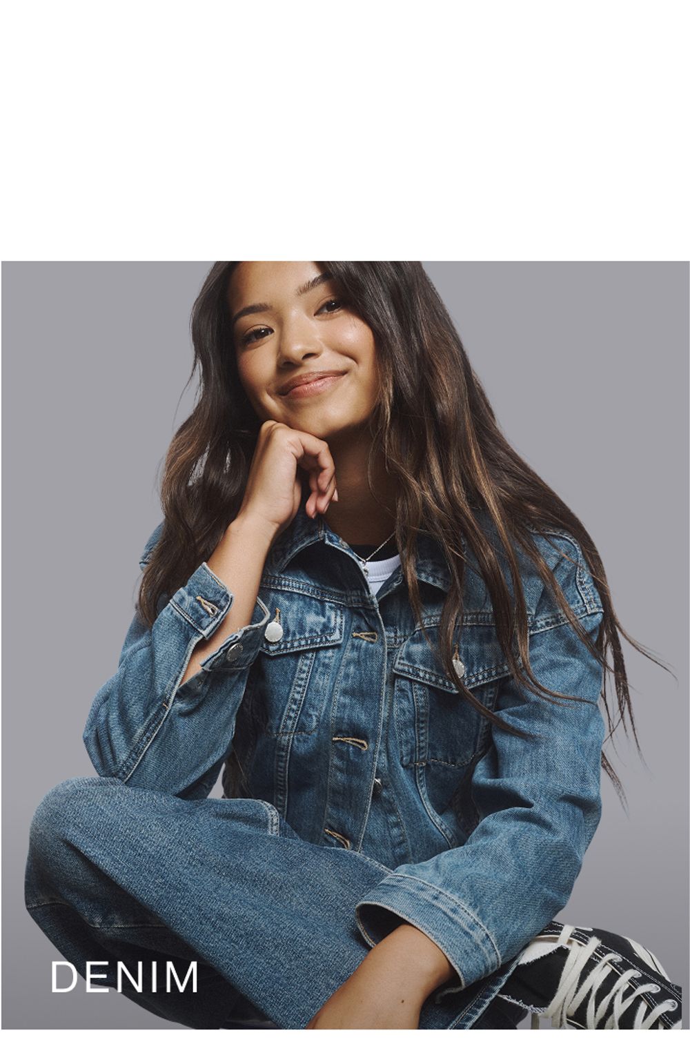 Young girl sitting on a chair with legs crossed wearing a denim set. SHOP TEEN DENIM.