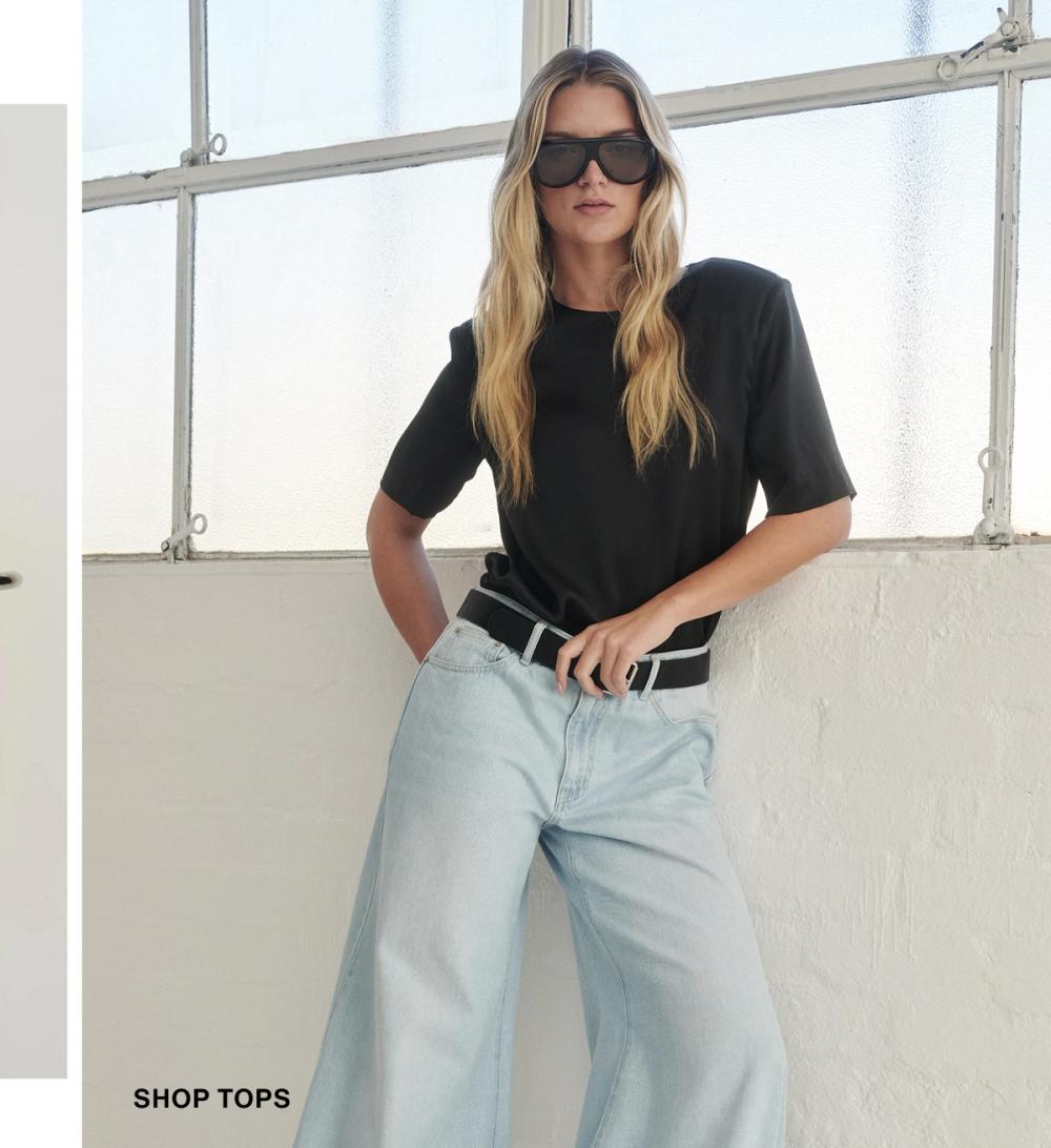 Model posing against a white wall wearing a black tee and jeans. SHOP TOPS.