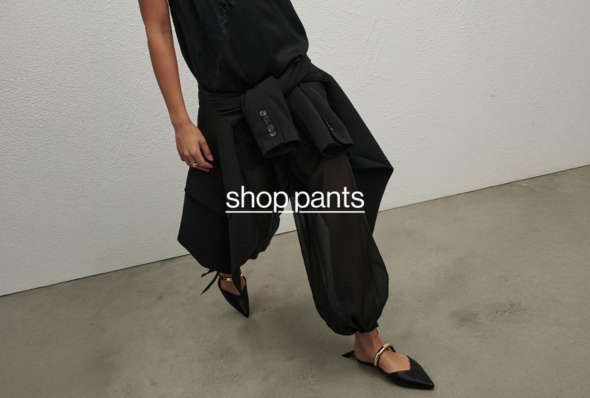 Model in relaxed black pants with a black sweater tied at the waist, walking across a concrete floor.