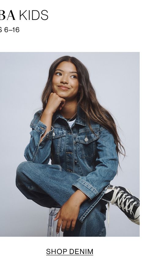 Young girl sitting cross-legged wearing a denim jacket, jeans, and sneakers