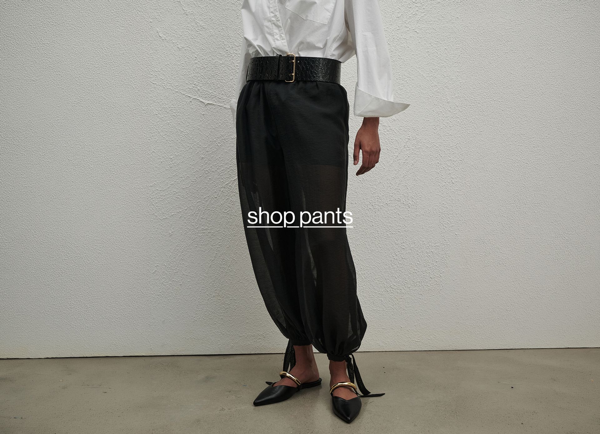 Model in relaxed black pants with a black sweater tied at the waist, walking across a concrete floor.