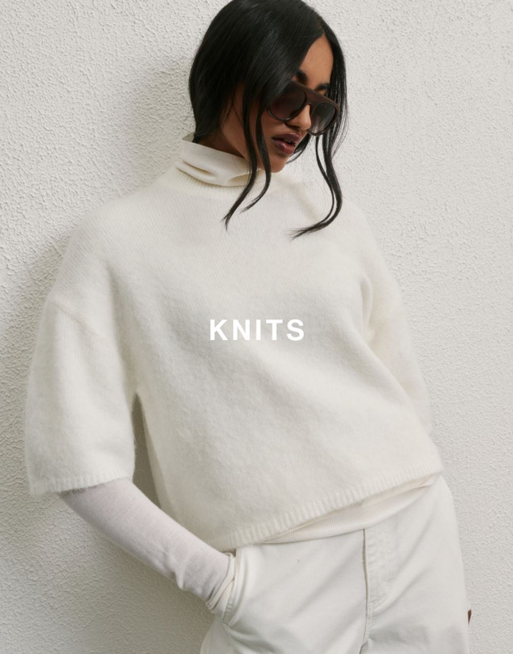 Model wearing a striped knit sweater and holding a brown woven bag, posed against a neutral background. Shop knits.