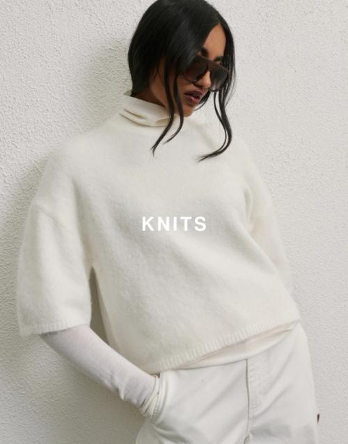 Model wearing a striped knit sweater and holding a brown woven bag, posed against a neutral background. Shop knits.