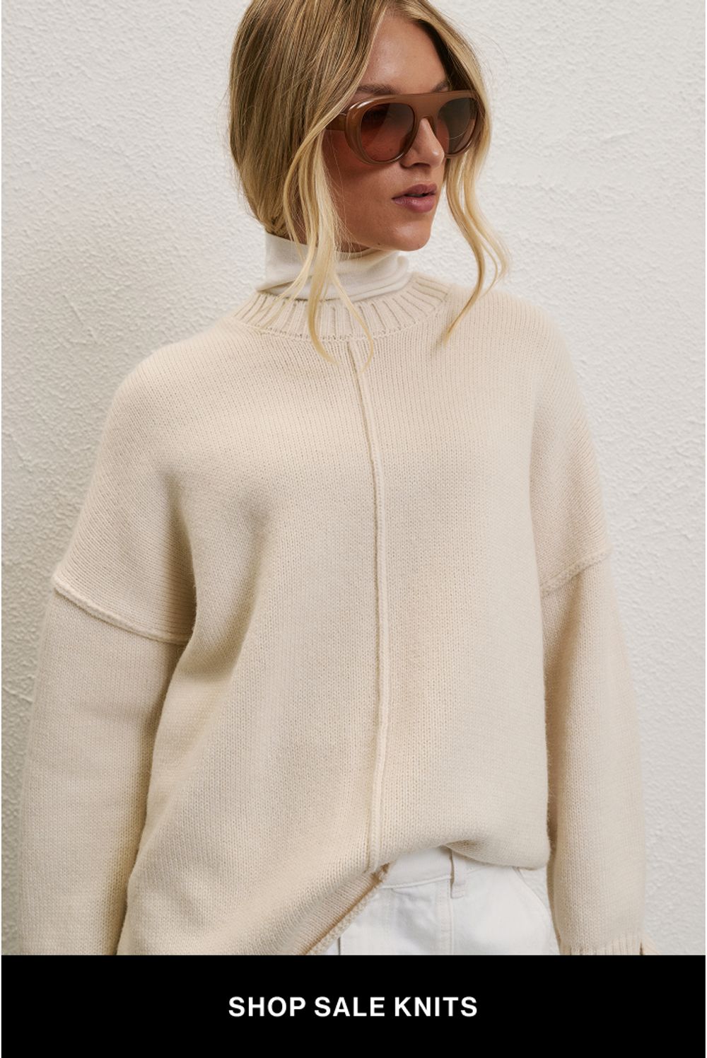 A model wearing a cream knit against a neutral background. SHOP SALE KNITS.