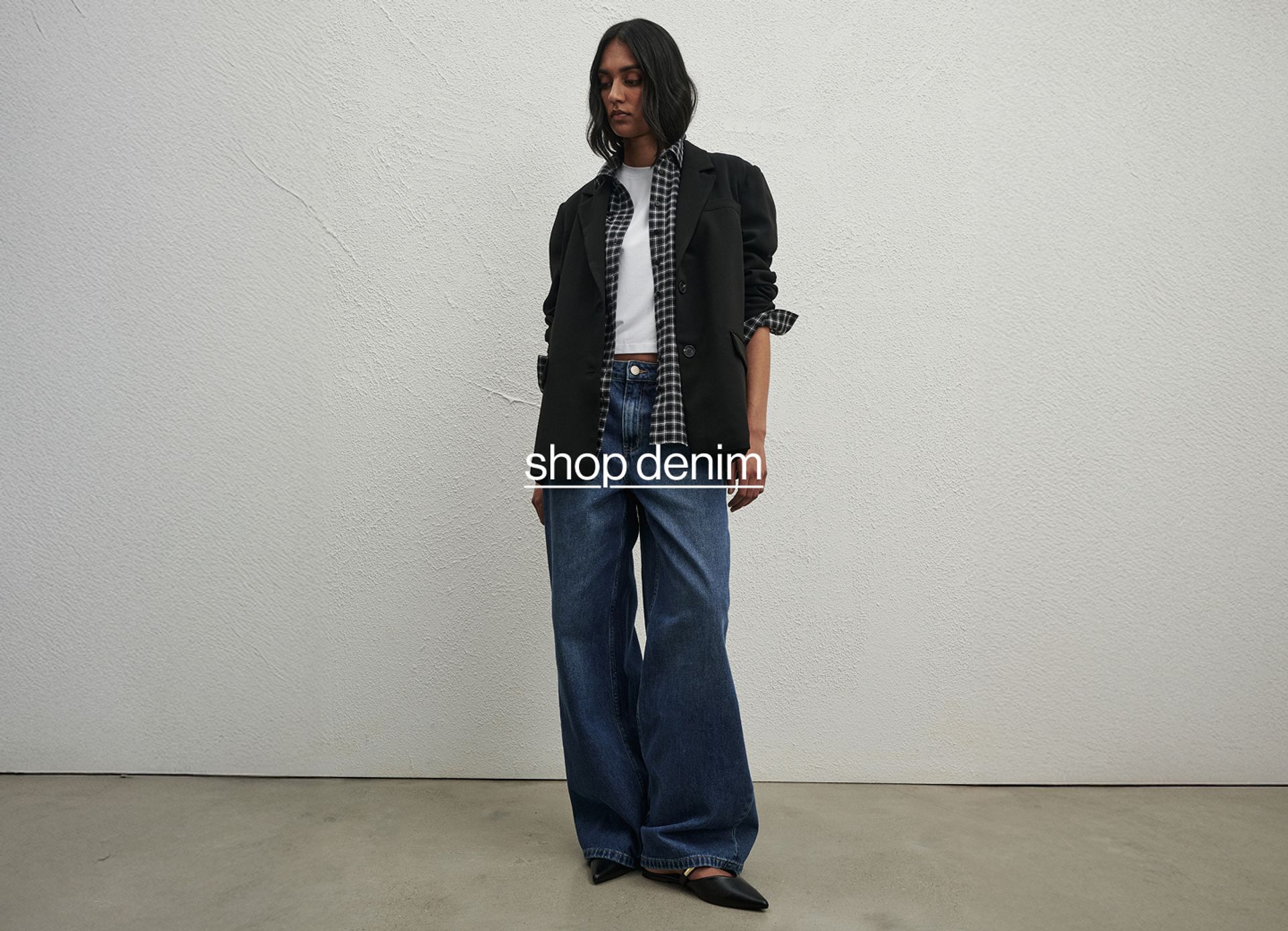 Model wearing a black jacket and wide-leg blue jeans, standing against a light concrete wall.