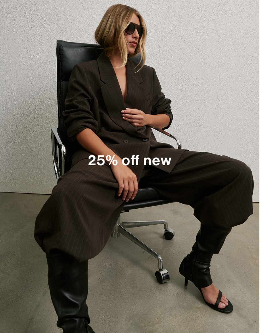Woman in oversized brown suit sitting in office chair wearing sunglasses, promoting 25% off new arrivals.