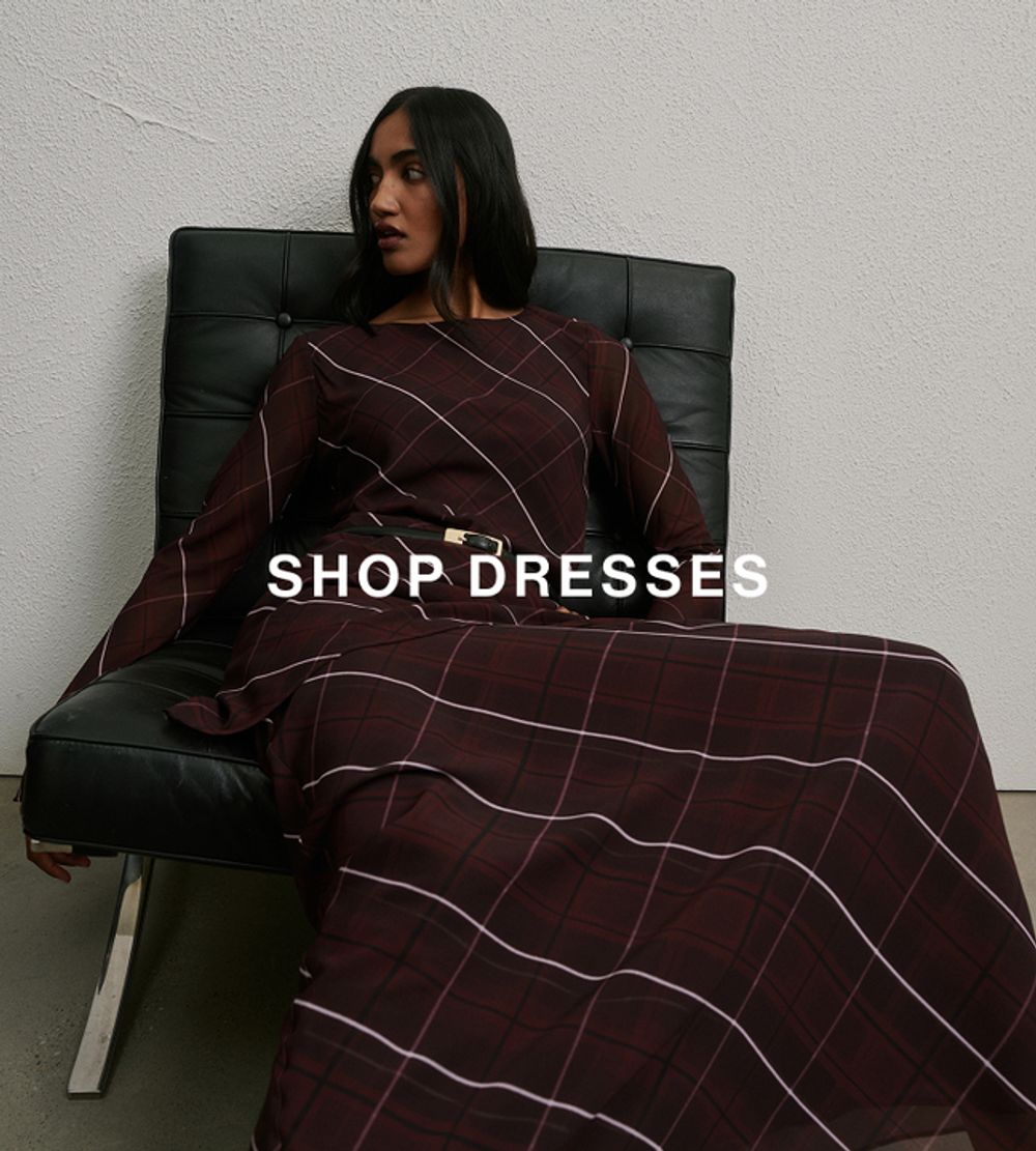 Woman posing in a modern chair wearing a checkered dress with text overlay that says "SHOP DRESSES"