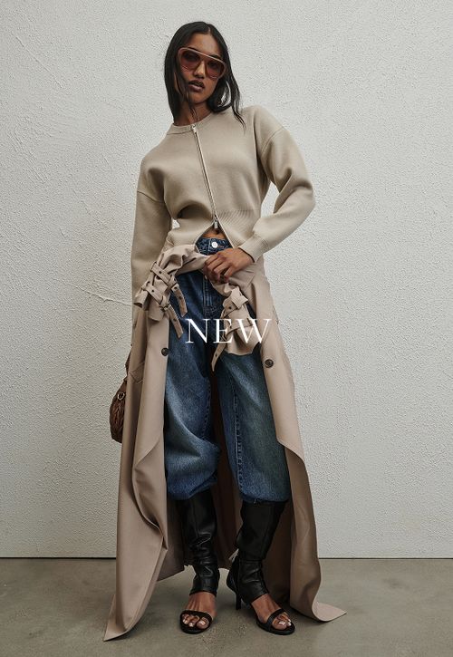 Woman standing in front of a white wall wearing a cream knit with text that reads "New"