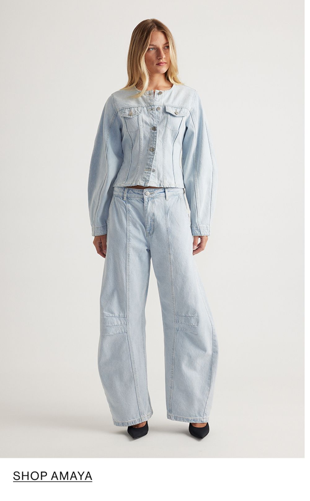 Woman standing in front of a white wall wearing a matching denim jacket and jeans set with texts that reads shop Amaya