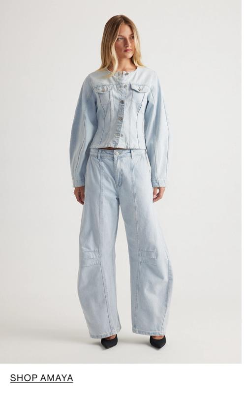 Woman standing in front of a white wall wearing a matching denim jacket and jeans set with texts that reads shop Amaya