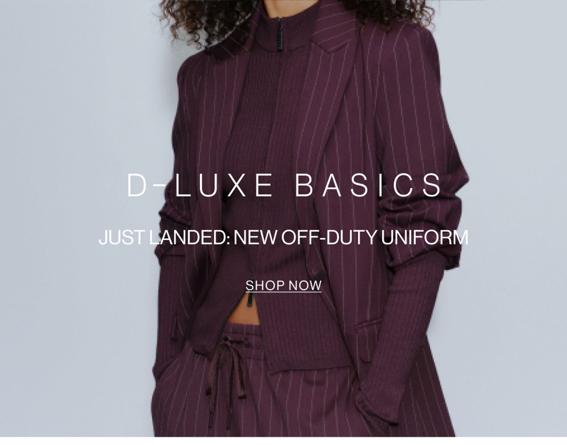 Model in a plum suit jacket standing in front of a white wall with text overlay which states "DLUXE BASICS"