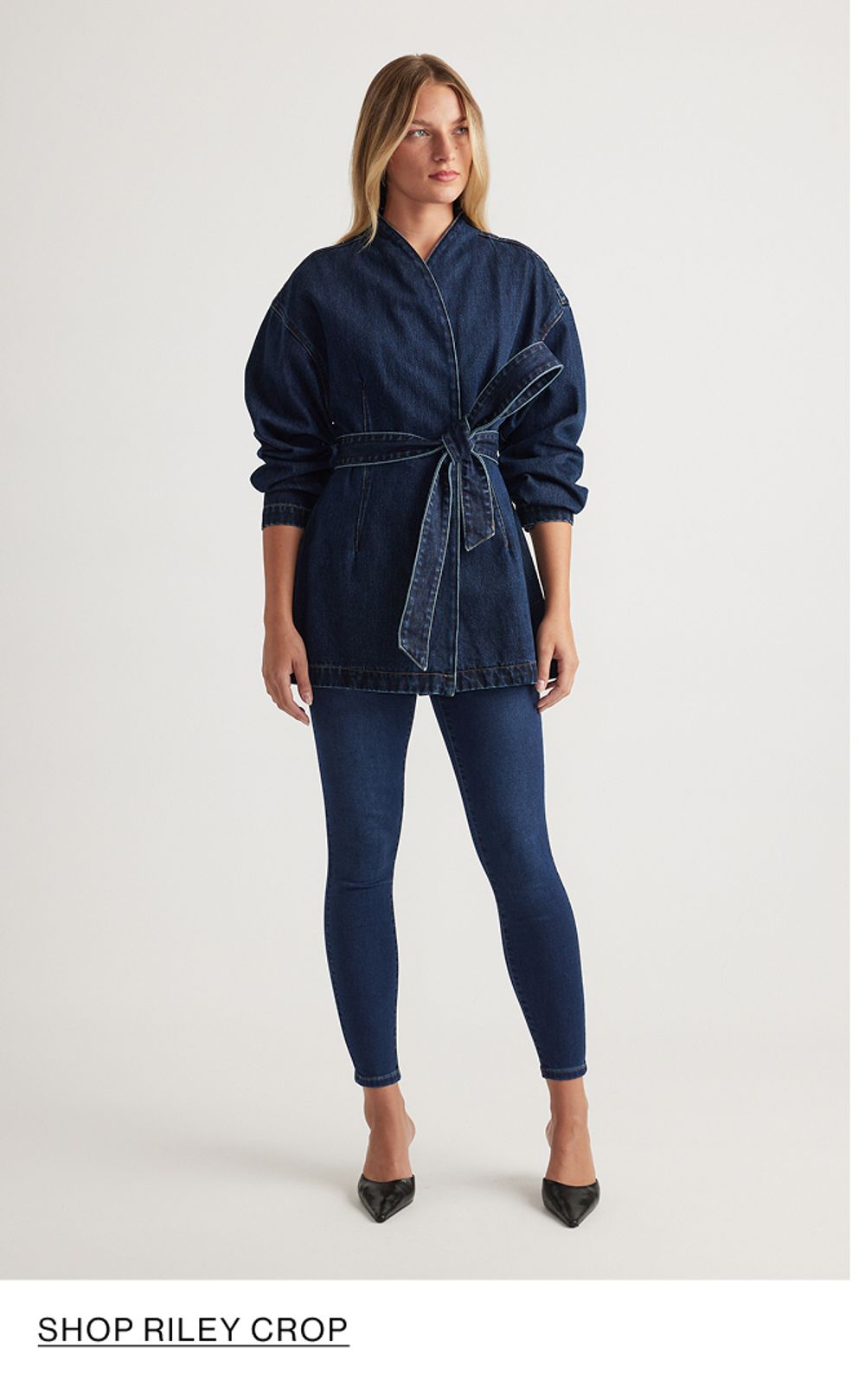 Woman standing in front of a white wall wearing a matching denim jacket and jeans set with texts that reads shop Crop Riley