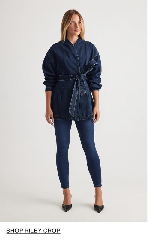 Woman standing in front of a white wall wearing a matching denim jacket and jeans set with texts that reads shop Crop Riley