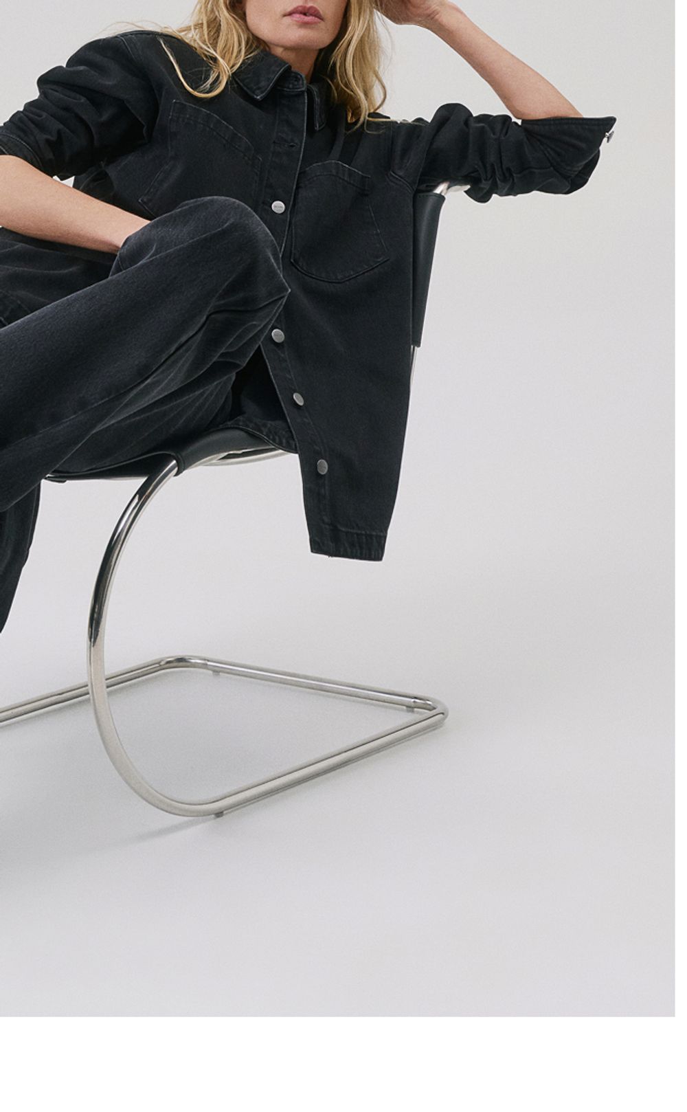 Woman sitting in a modern chair with her legs crossed and leaning to the side, wearing a black denim jacket and jeans set with black pointed heels.