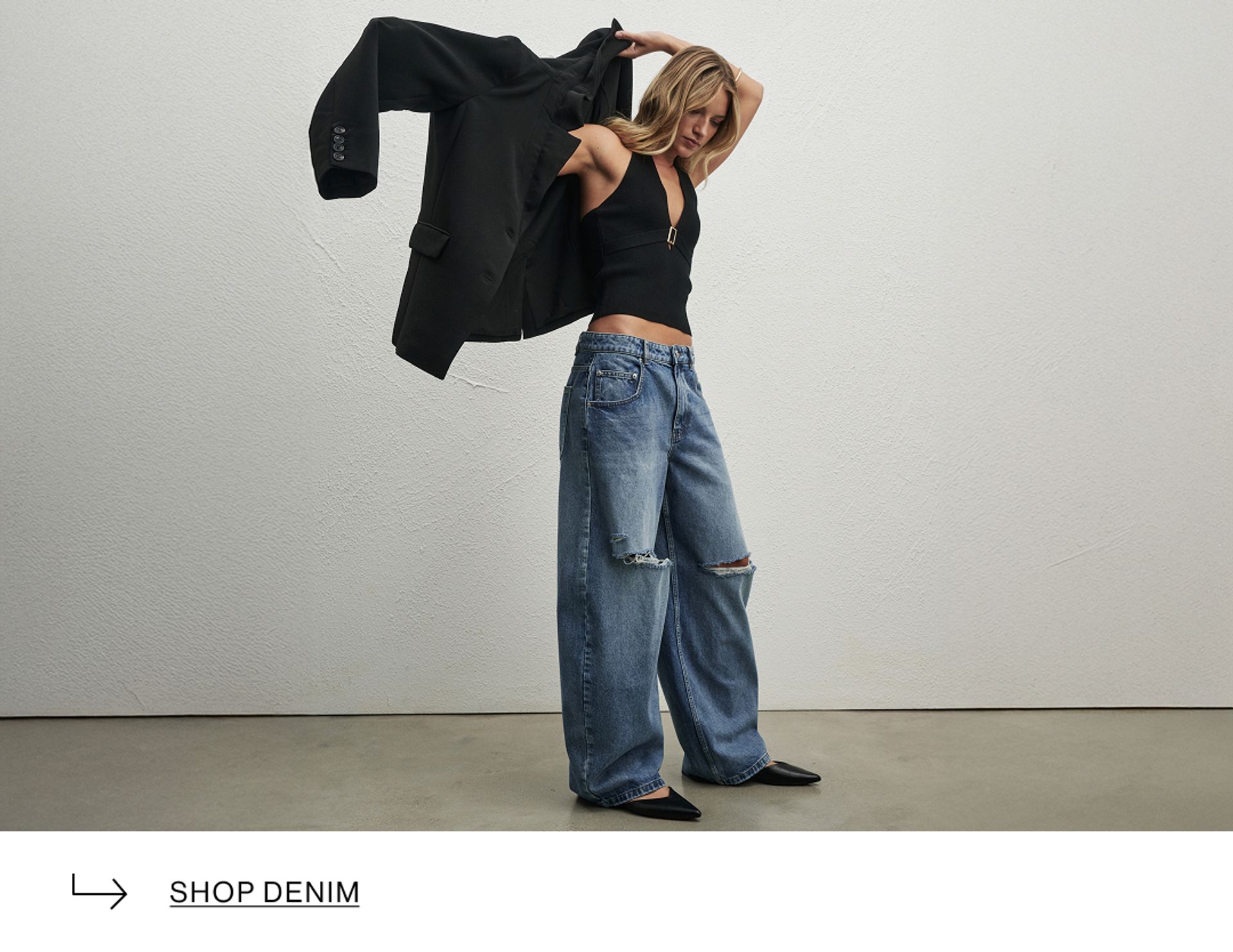 Model putting on a black jacket, wearing denim ripped jeans, standing against a minimalist white wall and gazing downward.
