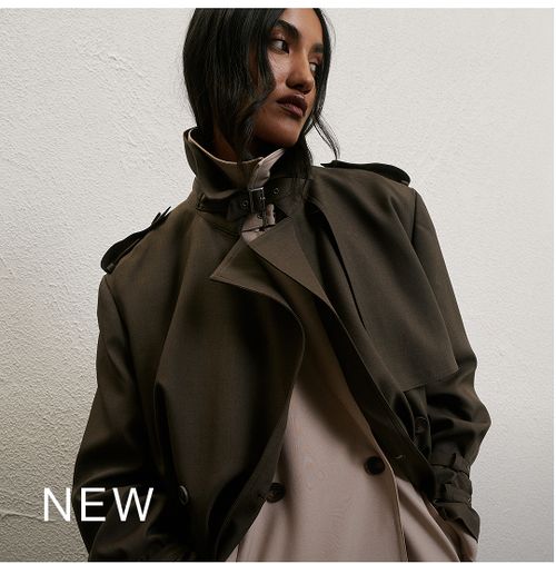 Woman standing in front of a white wall wearing a khaki trench coat with text that reads "New"