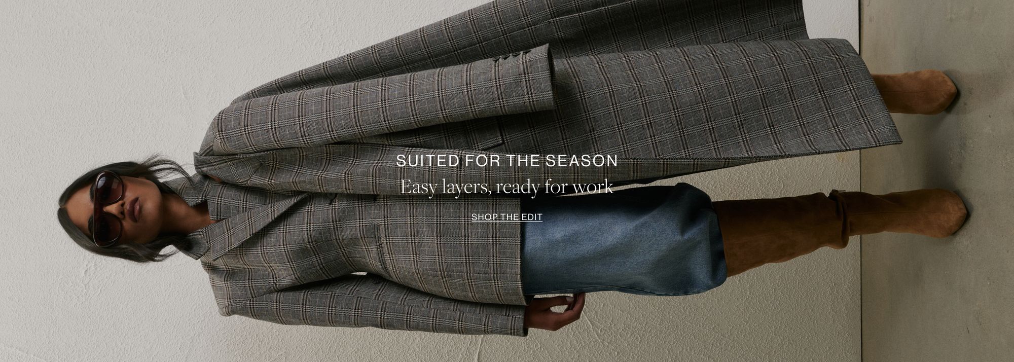 Woman standing in front of a white wall, wearing sunglasses and a long checkered coat with text that reads "Suited for the season. Easy layers, ready for work. Shop the Edit"