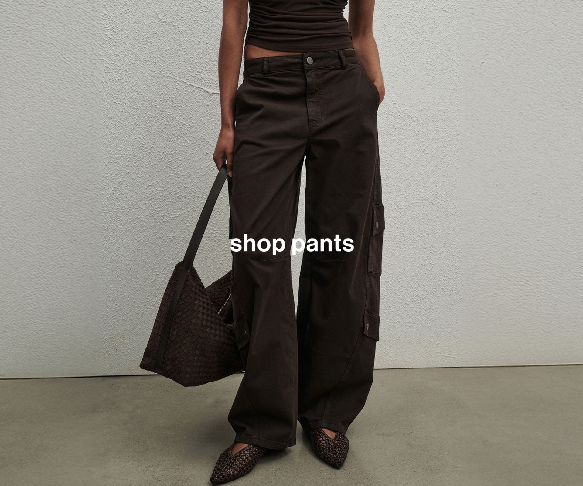 Woman wearing brown cargo pants and carrying a brown bag, standing in front of a white wall with one hand in her pocket.