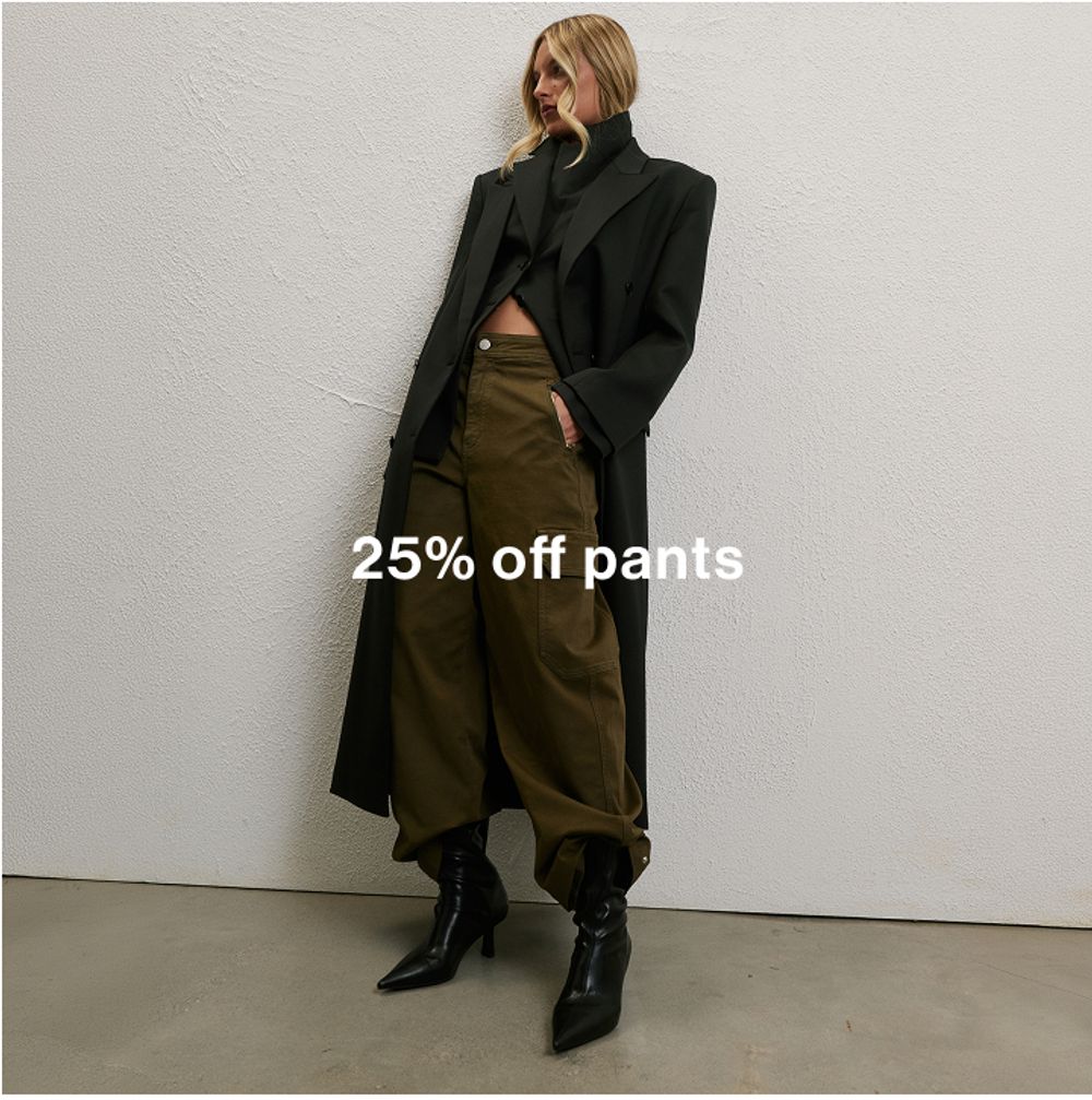 Woman wearing a long black coat, green pants and boots standing in front of a white wall with text that states 25% off pants