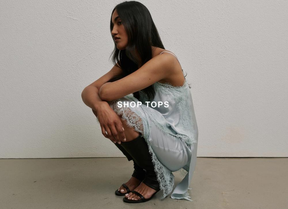 Woman posing in front of a white wall, crouching down wearing a matching blue lace set with text that reads "shop tops" 
