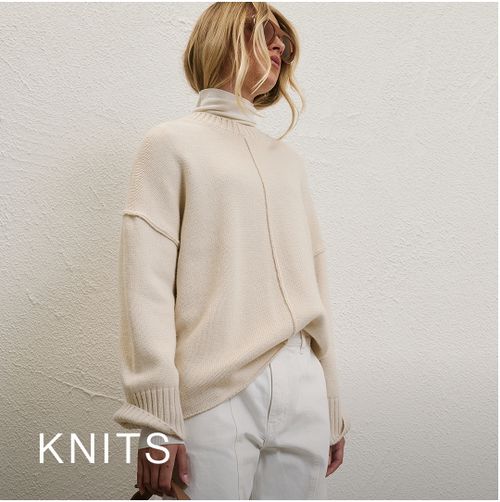 Woman posing in front of a white wall wearing a cream long sleeve knit with text that states "Knits"