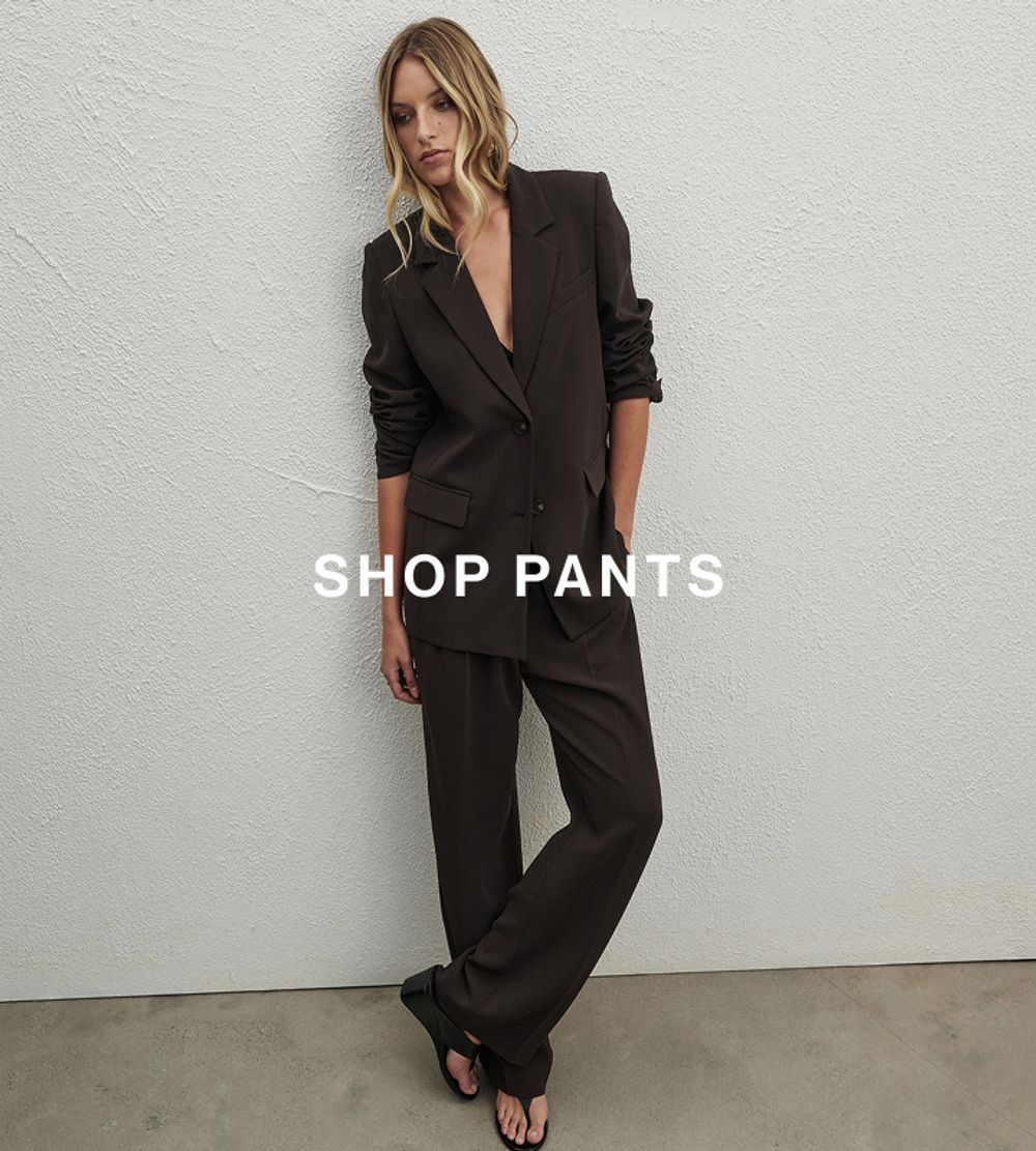 Woman posing in front of a white wall with her hand in her pocket wearing a brown suit with text that reads "shop pants" 