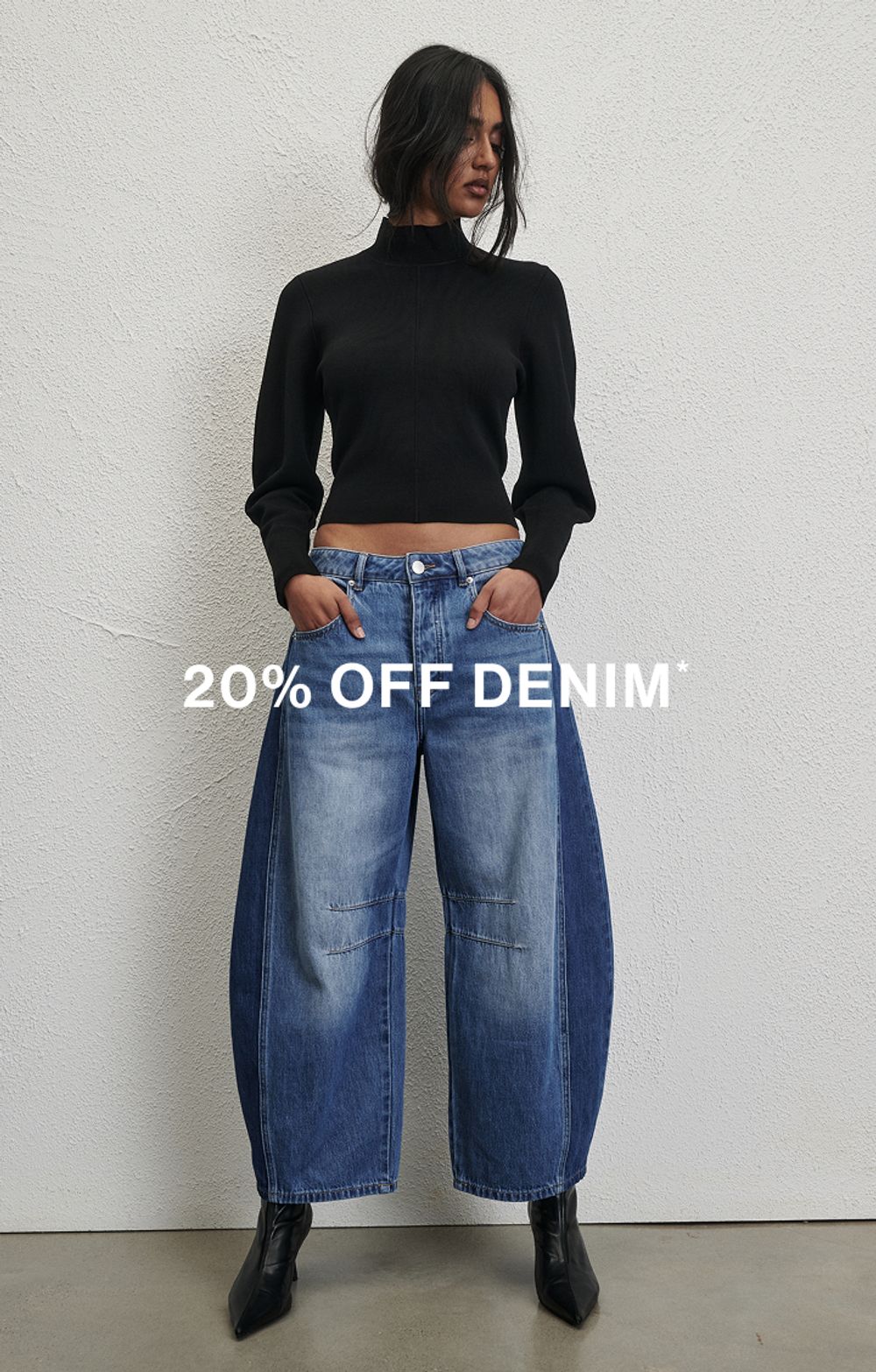 Woman posing in front of a white wall wearing a jeans and a black long sleeve top with text that states "20% off all denim"