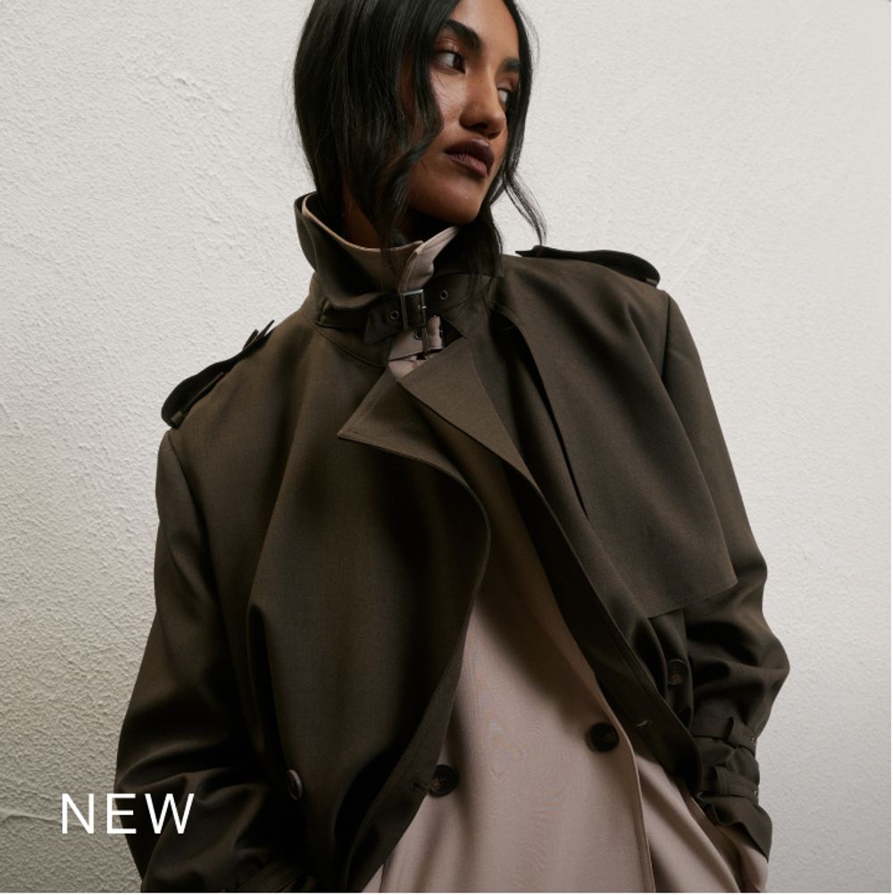 Woman standing in front of a white wall wearing a khaki trench coat with text that reads "New"