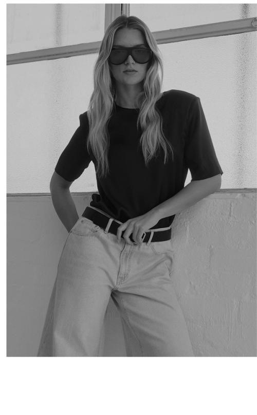 Woman standing in front of a white wall wearing a white shirt and wide jeans, with overlaid text that reads “Shop Elena.”