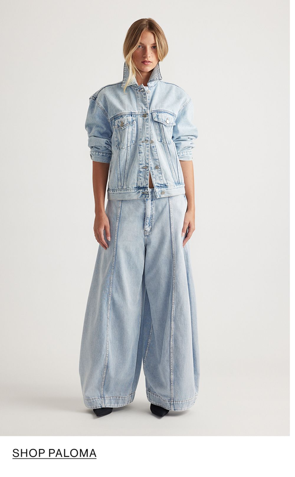 Woman standing in front of a white wall wearing a matching denim jacket and jeans set, with overlaid text that reads “Shop Paloma”