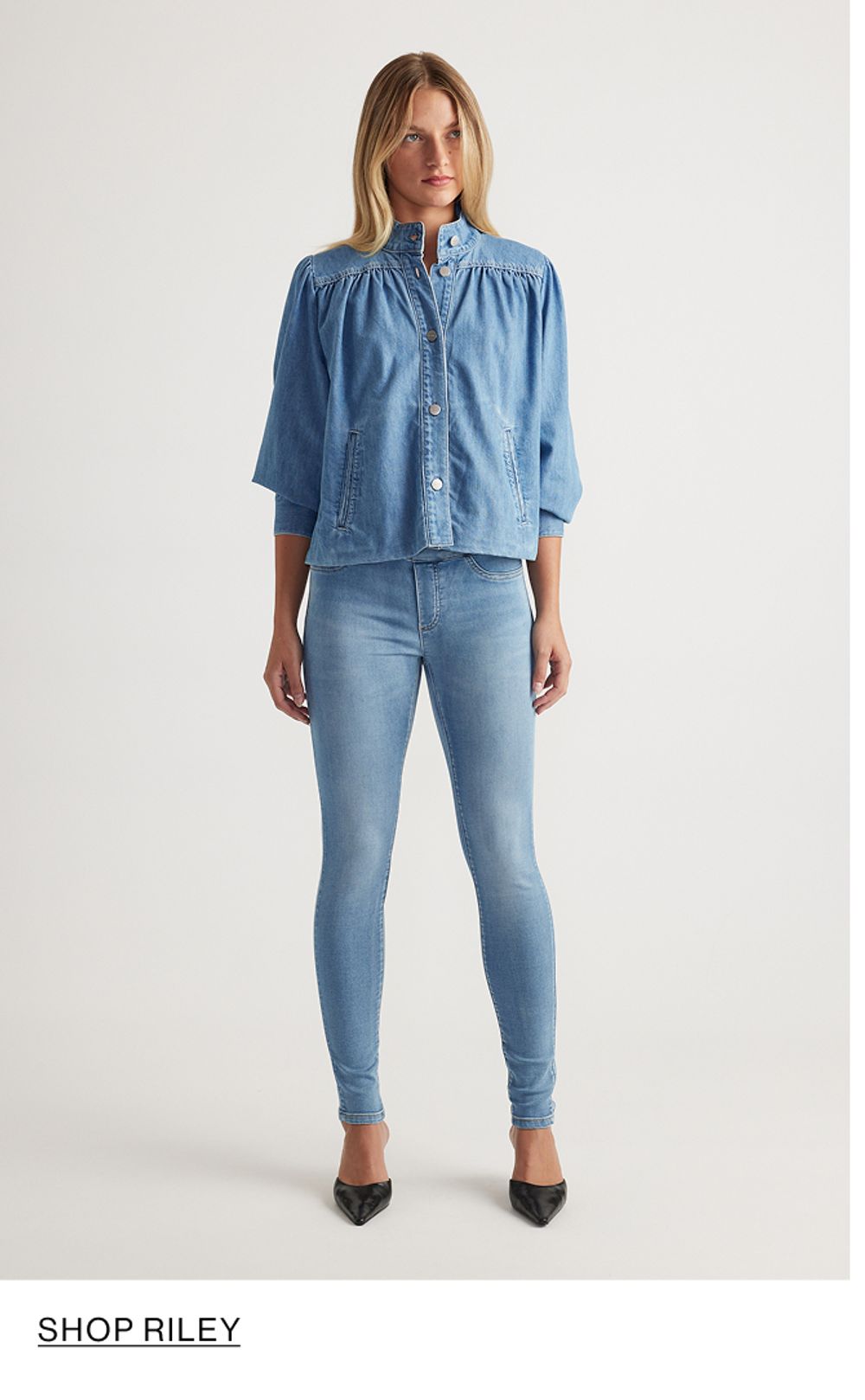 Woman standing in front of a white wall wearing a matching denim jacket and jeans set with texts that reads shop Riley